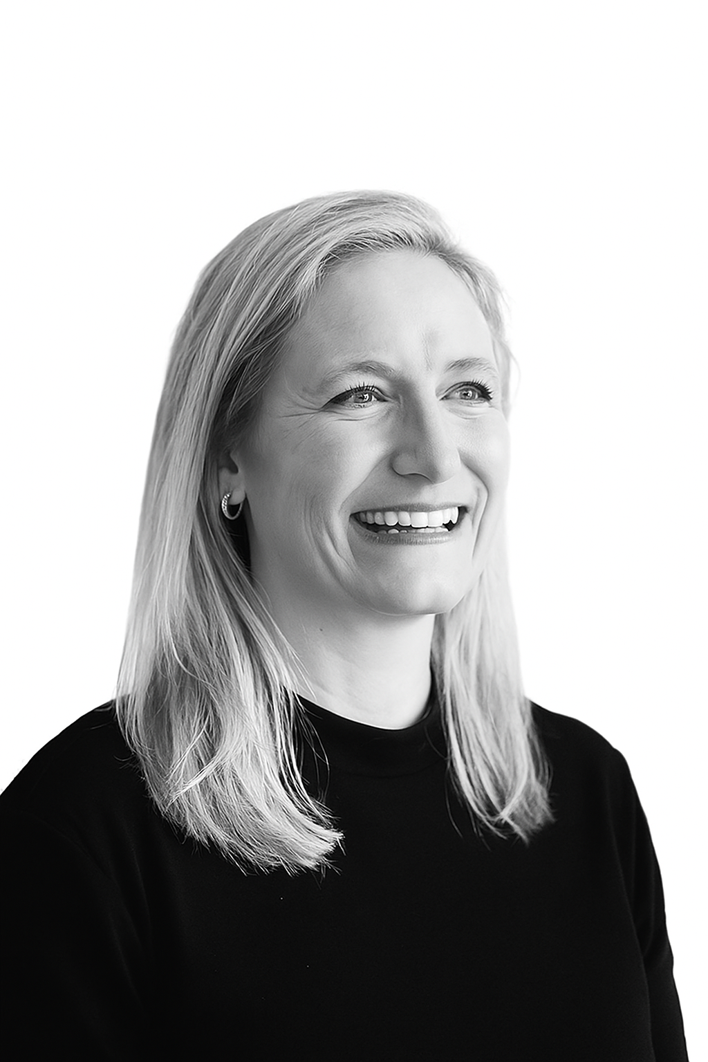 A black-and-white portrait of a smiling woman with shoulder-length blonde hair, wearing hoop earrings and a dark top.