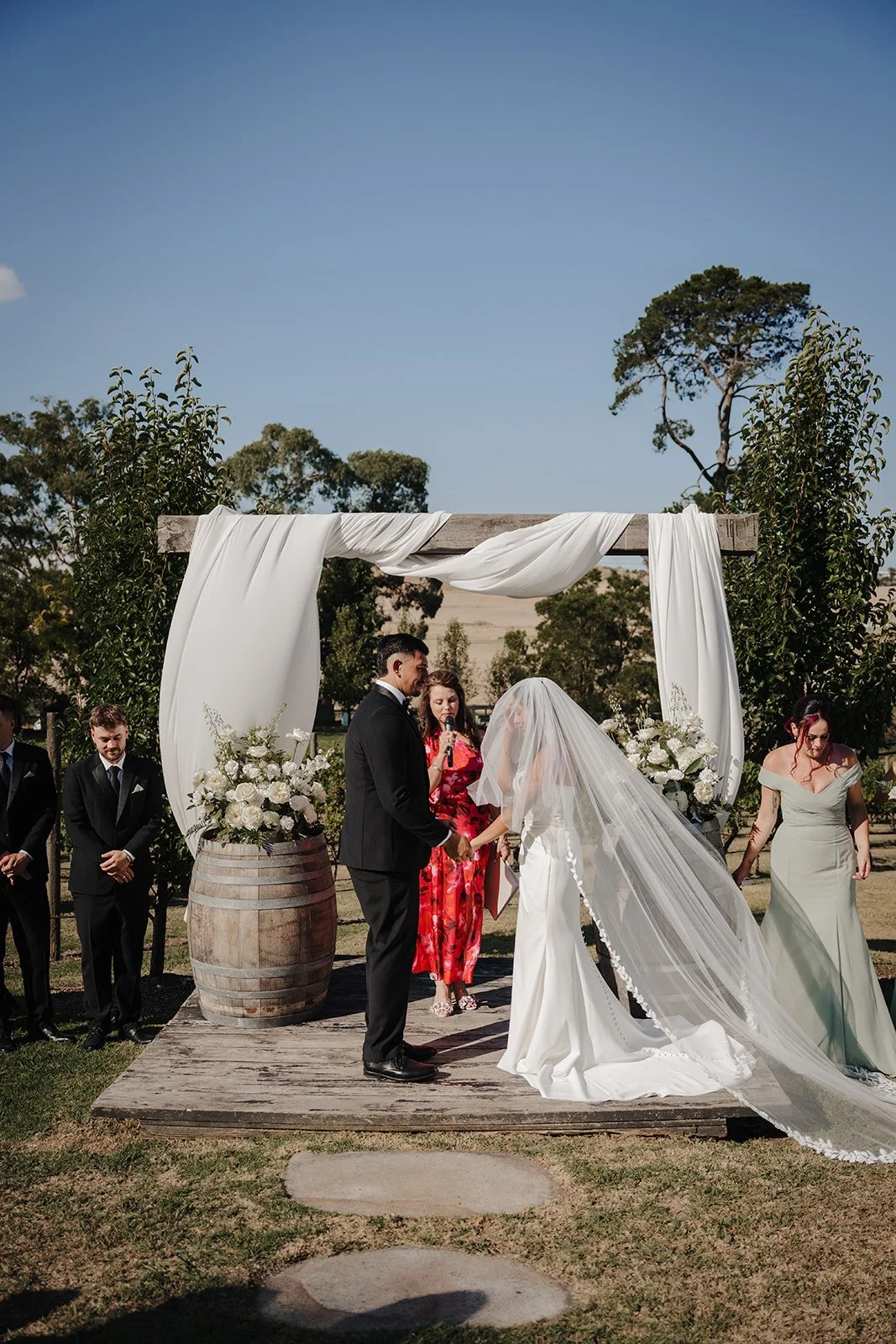 A wedding or formal ceremony with a woman in a gold and black dress and a man in a black suit shaking hands, with a woman in the background officiating and holding a microphone and paper, surrounded by white floral arrangements.