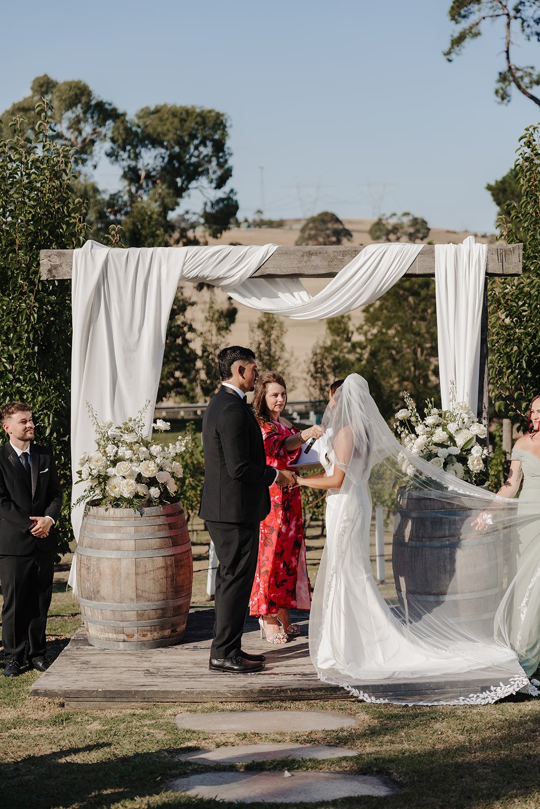 A wedding ceremony where a couple is exchanging vows in front of a woman with blonde hair, who is holding a book. The bride is wearing a black dress and the groom a black checkered suit. A woman in a black dress holds a bouquet on the left, and a man in a dark suit stands on the right. The backdrop has large white floral arrangements and a plain white wall, with seated guests watching.