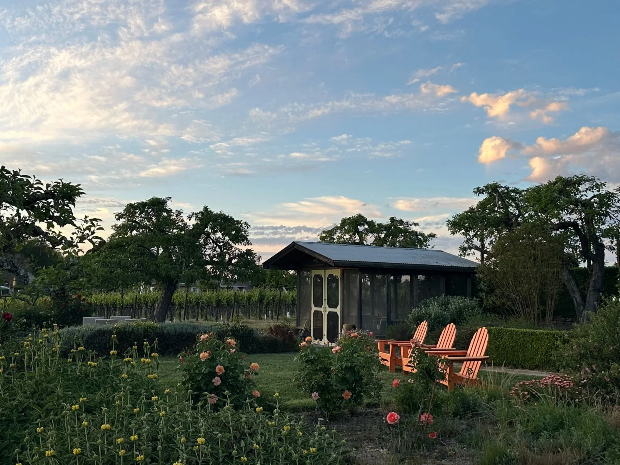 A garden with blooming pink and yellow flowers, surrounded by green trees and shrubs. There are four orange wooden chairs facing a small covered sitting area with a roof, set against a backdrop of a clear sky with scattered clouds.