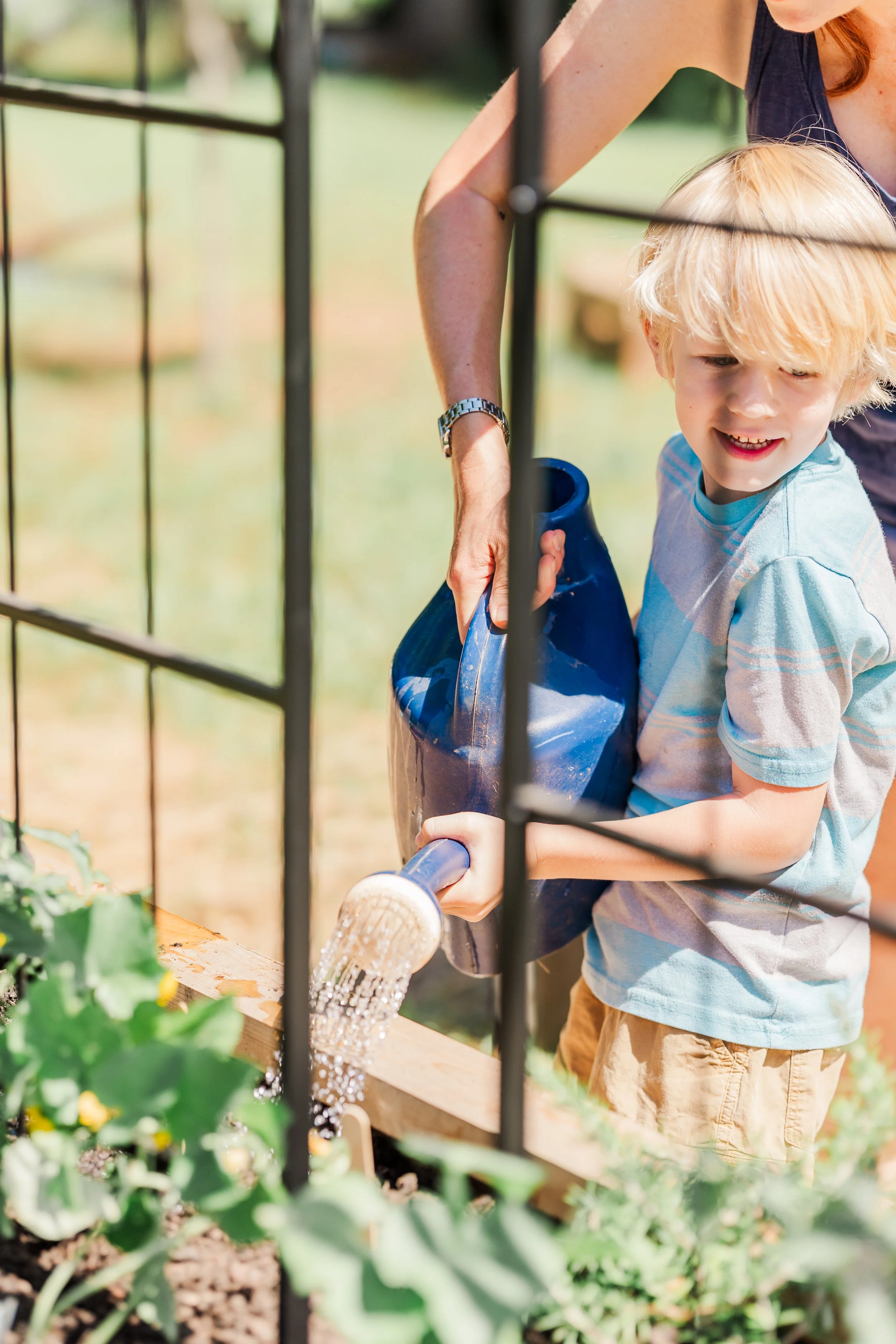 A woman helping a young boy water plants in a garden using a blue watering can.