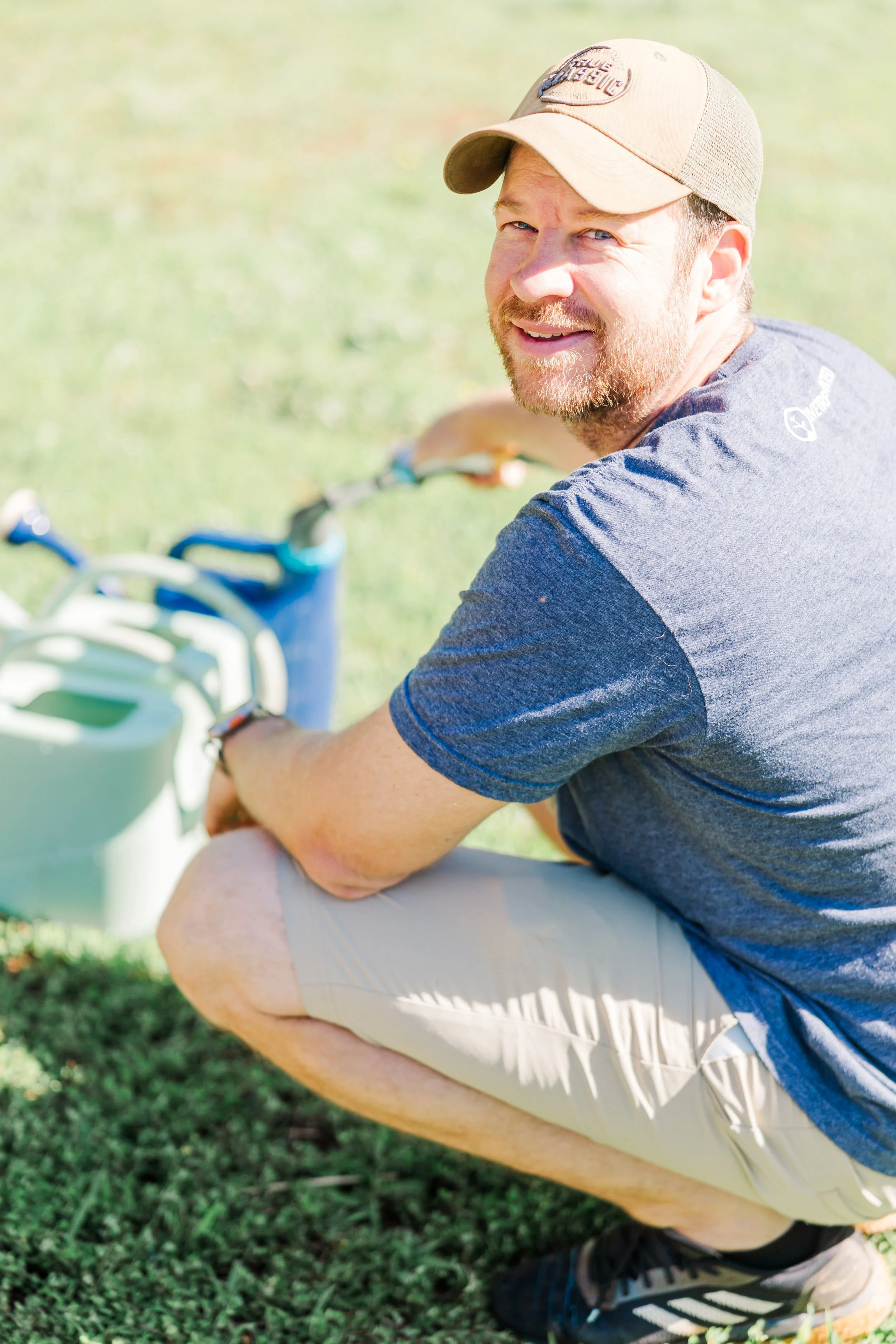 A man in a baseball cap, blue t-shirt, and khaki shorts squats on grass with a green water can and a blue water bottle in front of him, smiling at the camera.