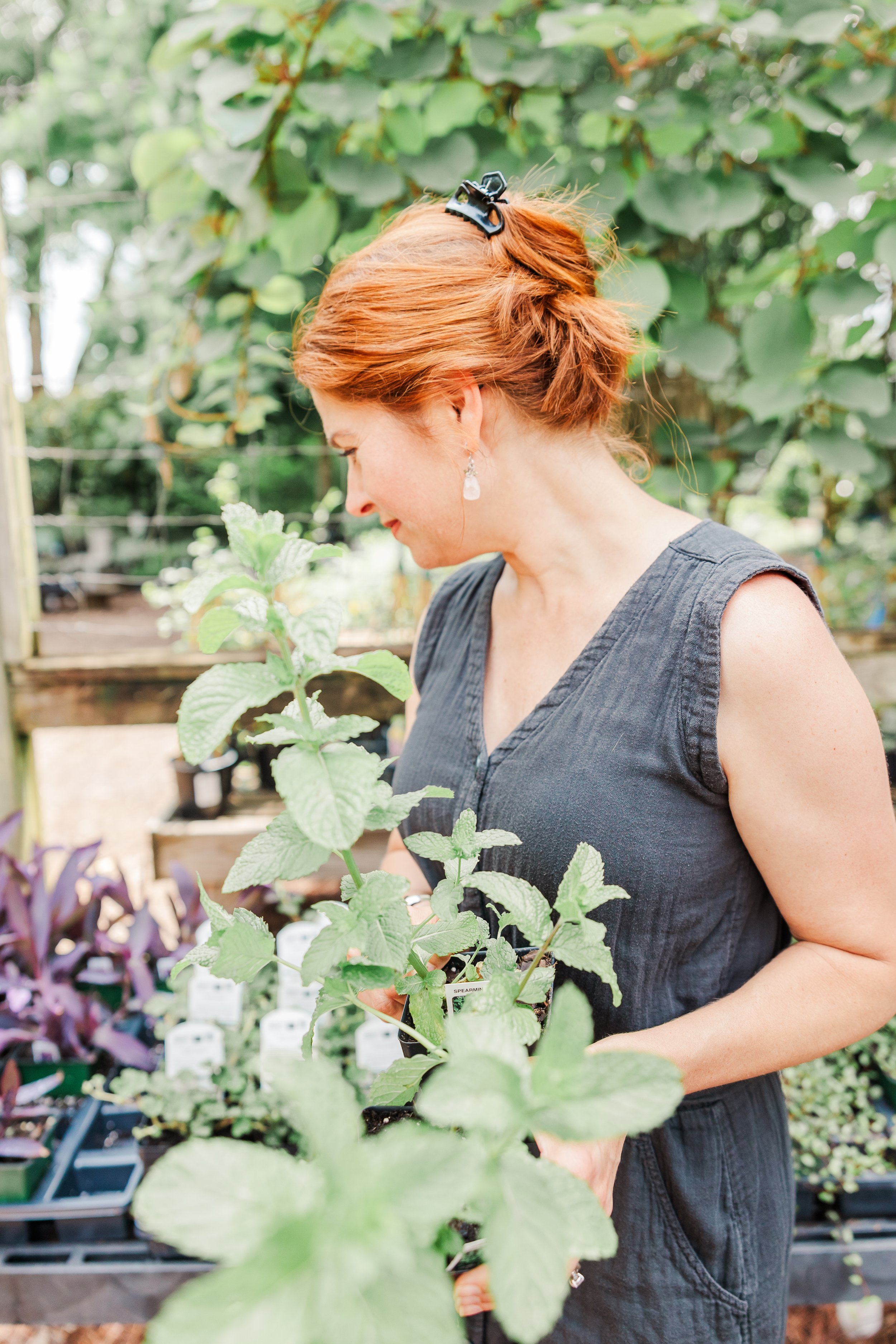 A woman with red hair, wearing a sleeveless black dress and earrings, is examining a potted plant in a garden center.