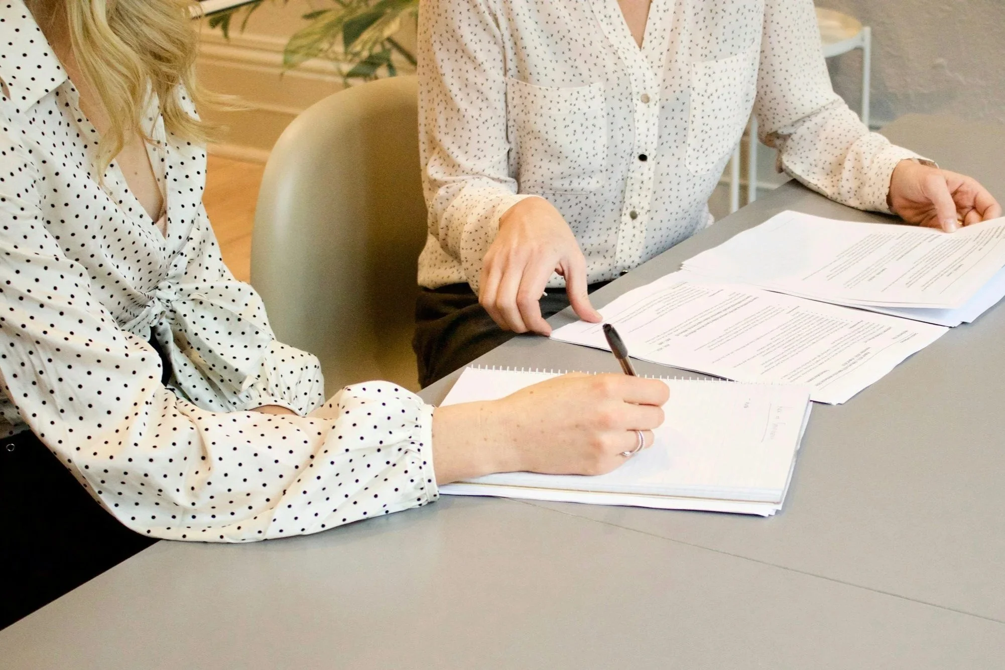 Two women sitting at a table, reviewing documents and taking notes, both wearing white blouses with black polka dots.