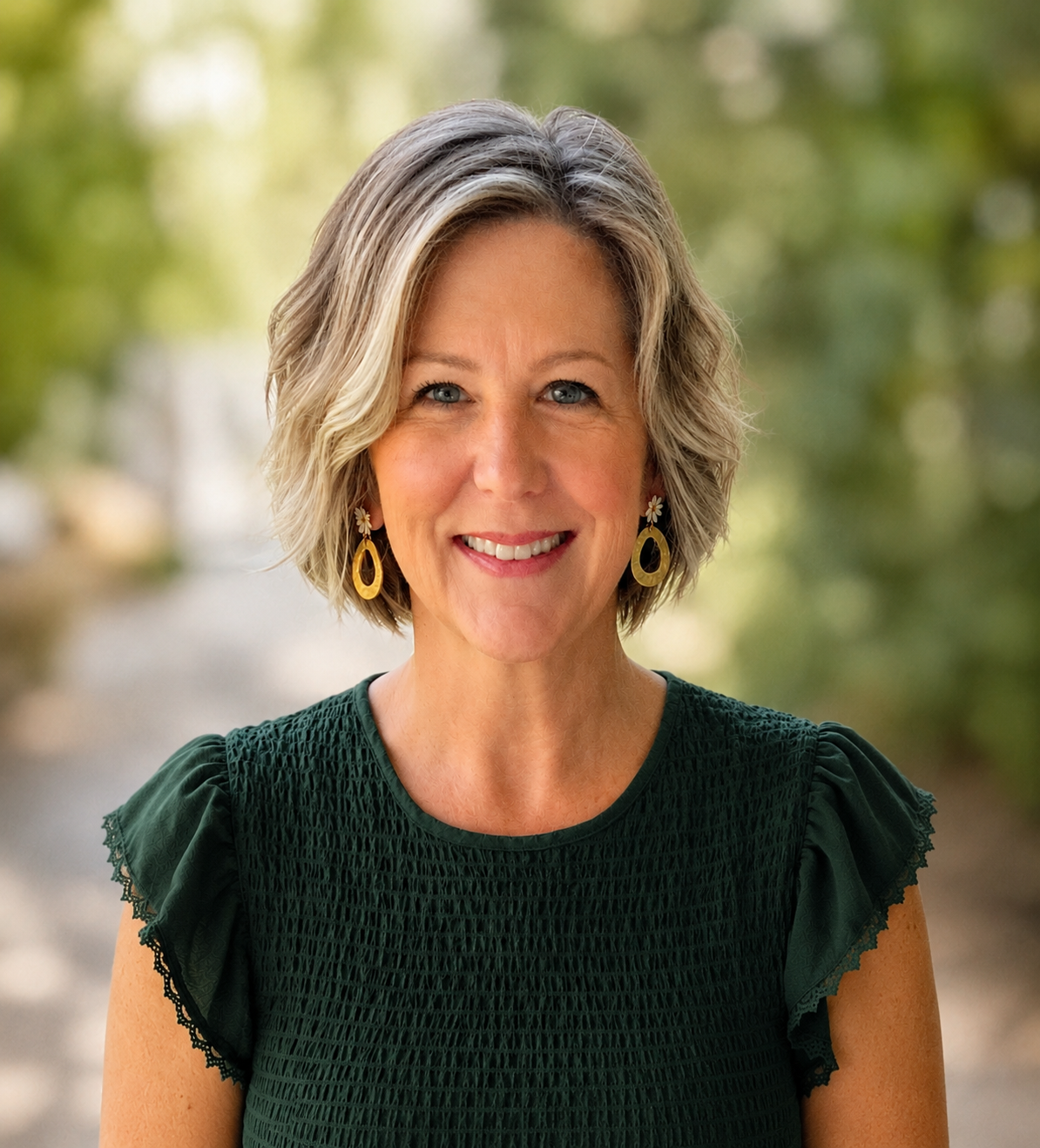 A smiling middle-aged woman with short, wavy blond hair and blue eyes, wearing a dark green top with ruffled sleeves and yellow earrings with daisy accents, outdoors with a blurred green and yellow background.
