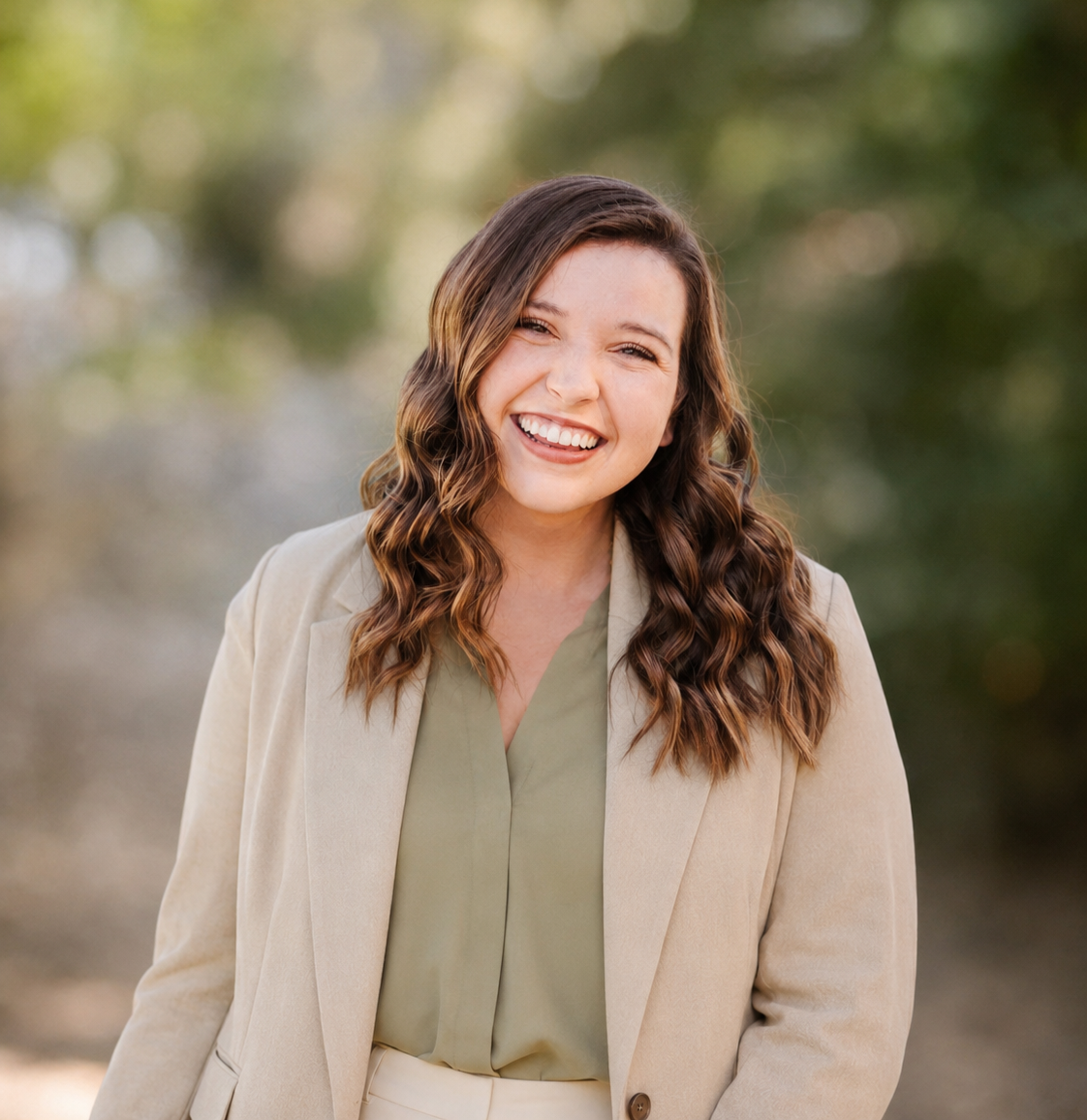 A smiling woman with long wavy brown hair, wearing a beige blazer and light green blouse, outdoors with blurred trees in the background.