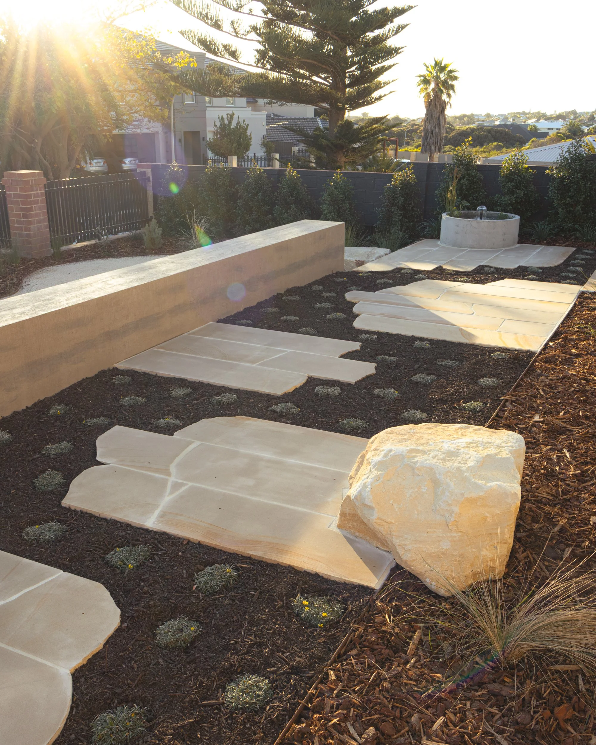 A garden yard with large cream-colored stepping stones on dark soil, a large rock, and a concrete water fountain in the background, illuminated by sunlight.