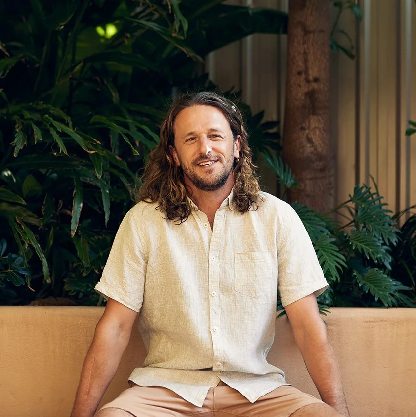A man with long, curly hair and a beard, wearing a short-sleeved beige shirt, sitting on a bench with lush green plants in the background.