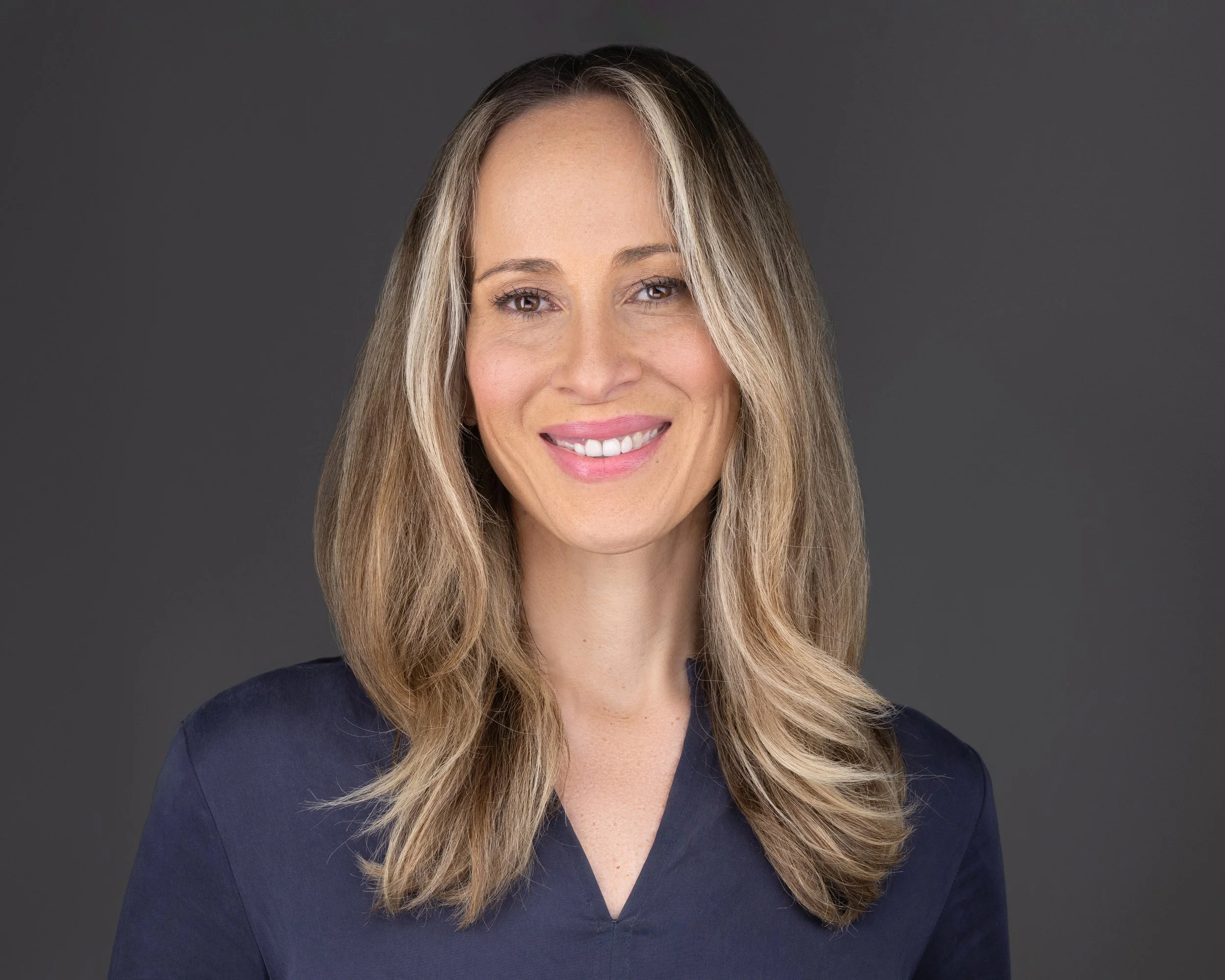 A smiling woman with long, wavy blonde hair, wearing a navy blue top, against a gray background.