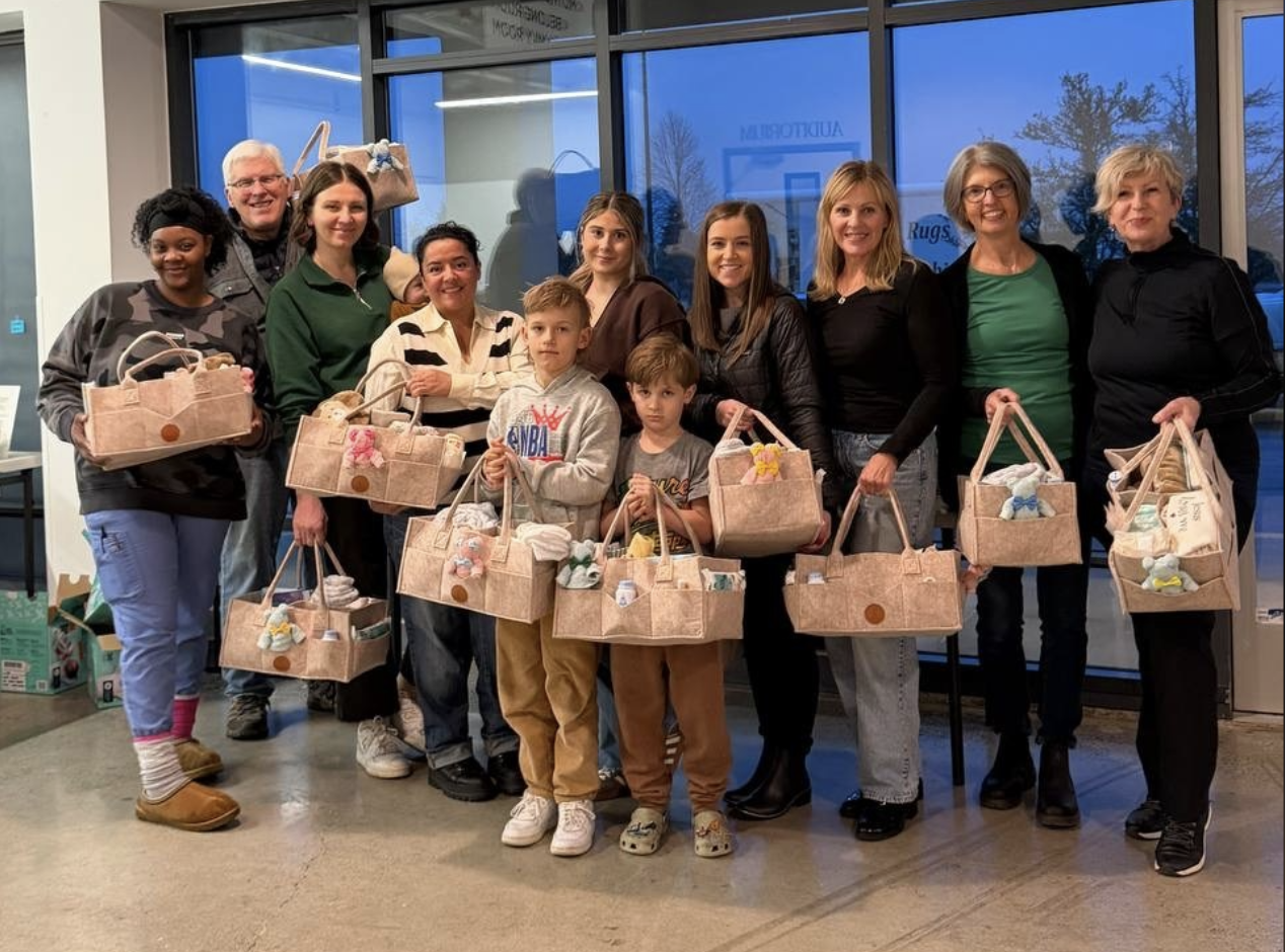 Group of eleven people, including children and adults, standing inside a building with large windows, holding beige baskets filled with items, smiling for the camera.