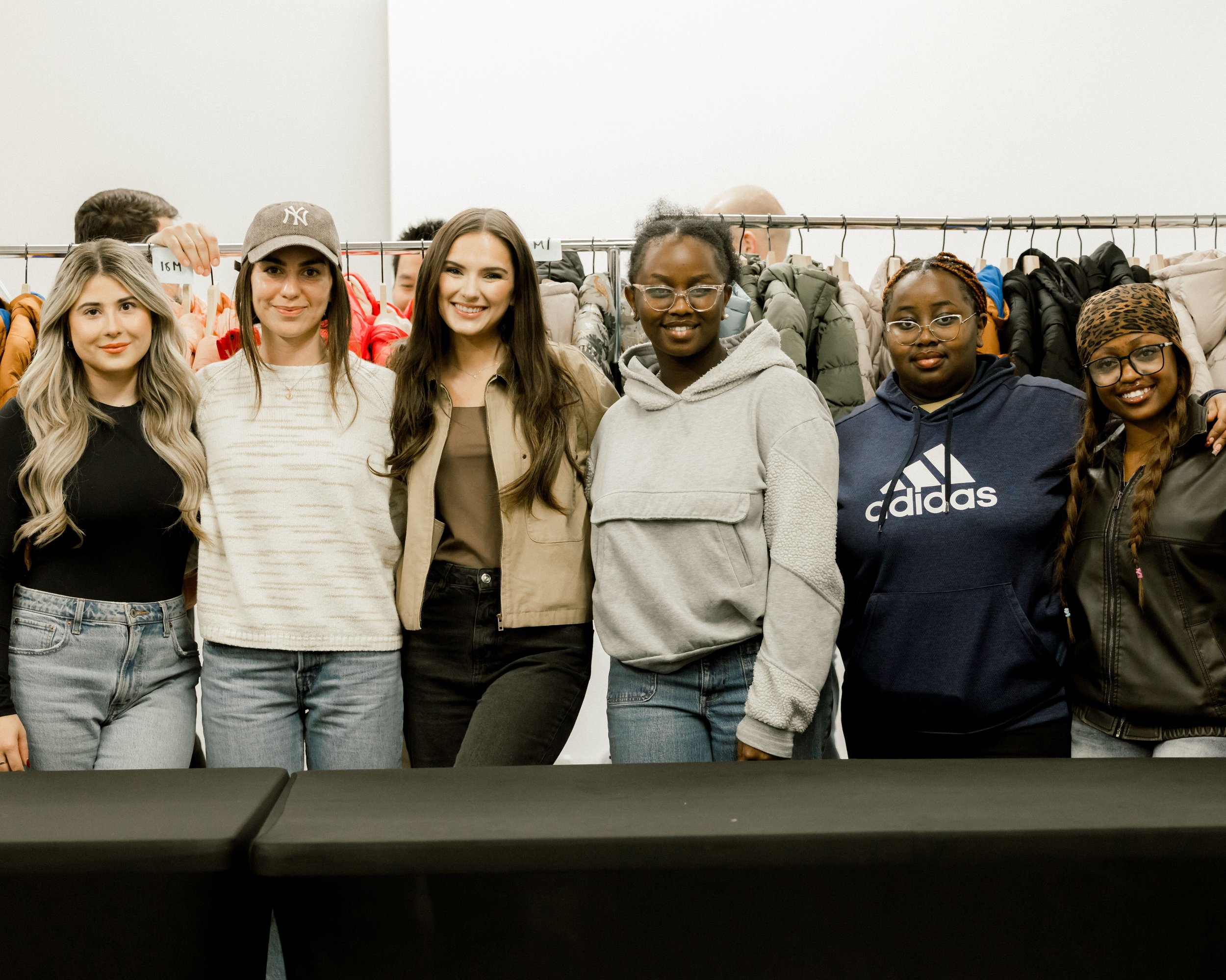 A group of six women standing together indoors, in front of a clothing rack with jackets, smiling at the camera.