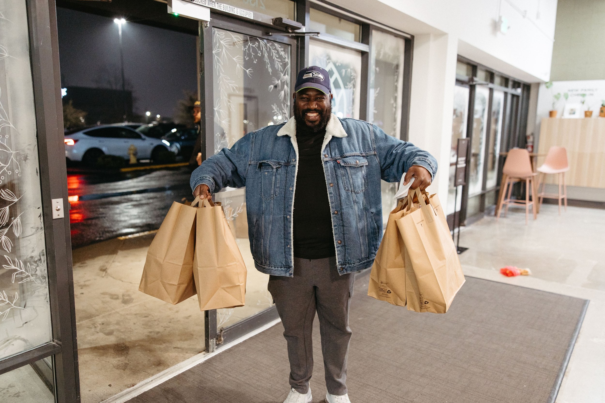 A man smiling outside a store at night, holding multiple paper shopping bags.