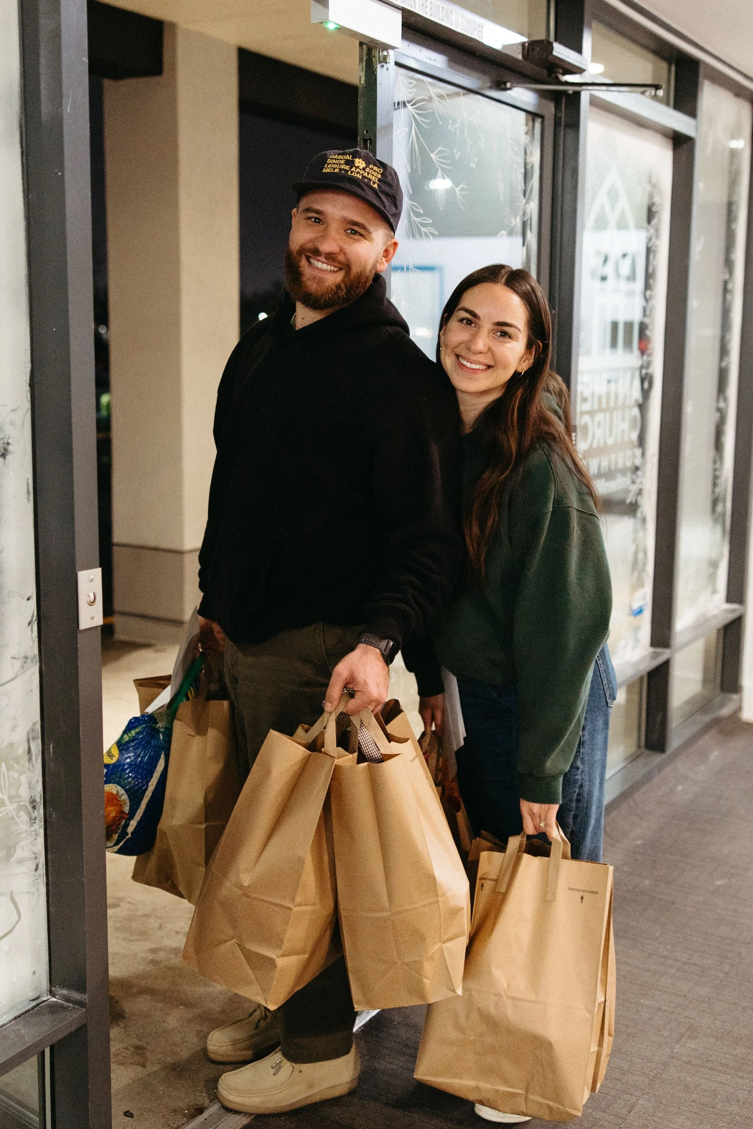 A happy young man and woman standing at a shopping mall entrance, holding multiple paper shopping bags, smiling at the camera.