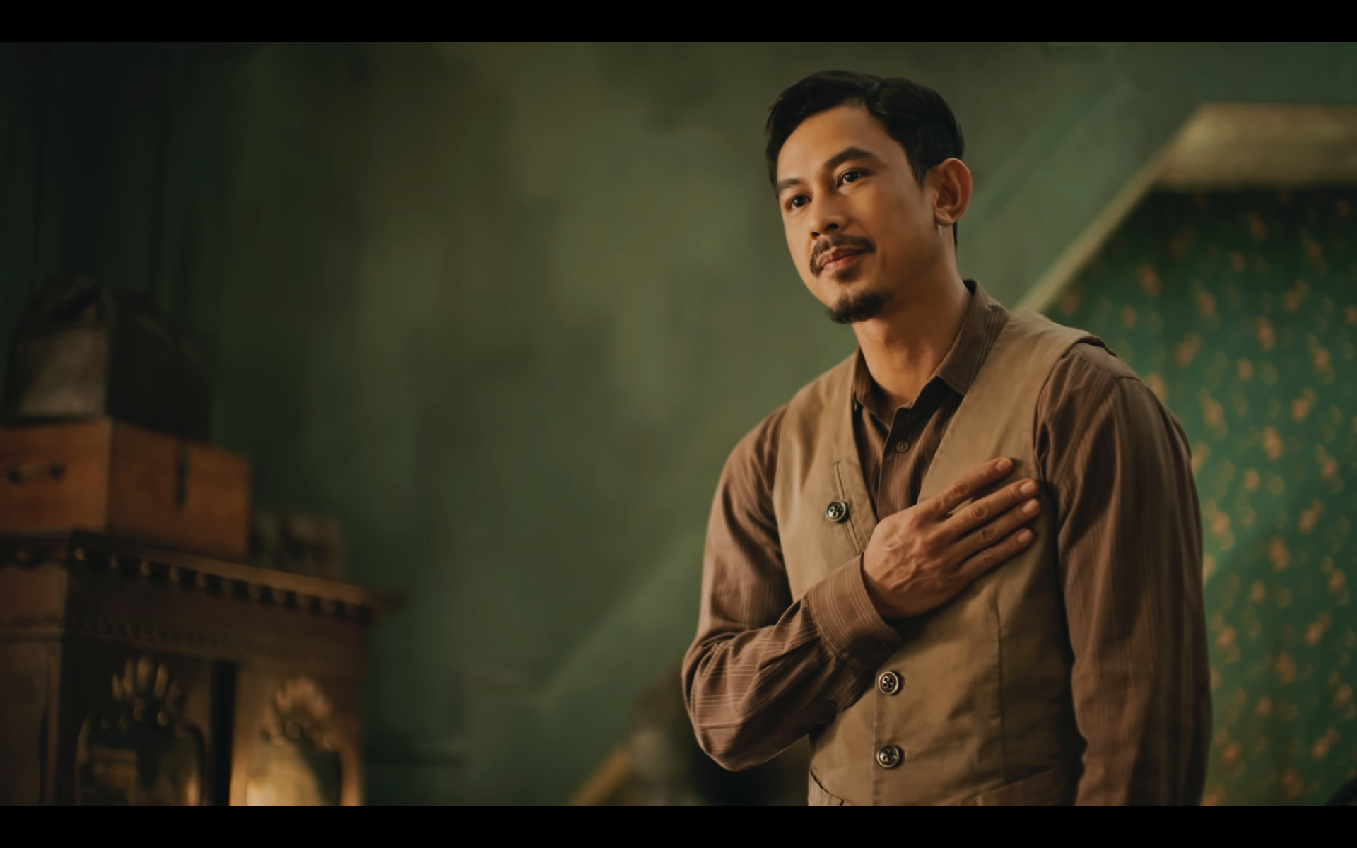 A man in period costume stands with his hand over his chest, looking thoughtfully to the side in an ornate room with green walls and a wooden cabinet.