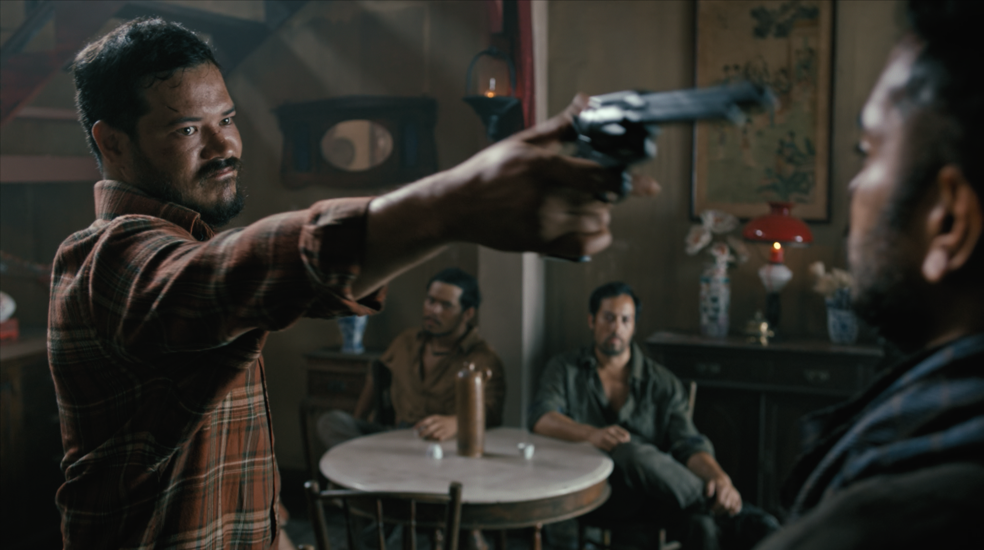 A man with a mustache and beard is pointing a handgun at another man inside a dimly lit room, while other men sit at a table in the background.