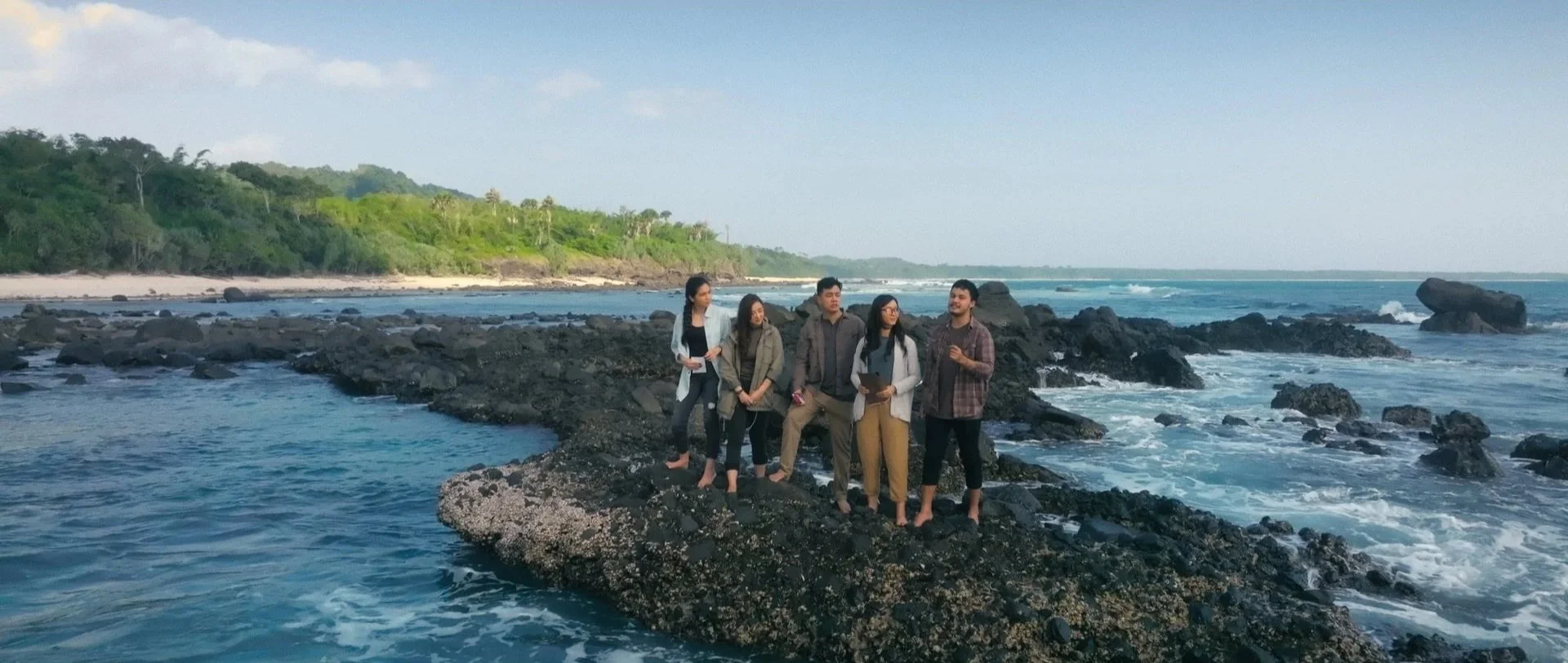 Group of people standing on rocks by the ocean at a scenic coastal location with lush green hills in the background.