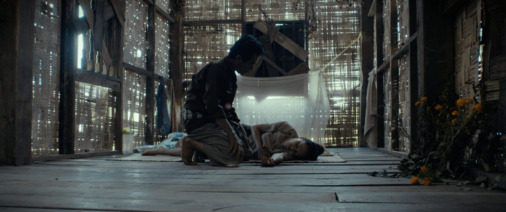 A man appears to be mourning over a young girl lying on the floor inside a rustic wooden house with sunlight filtering through gaps in the walls.