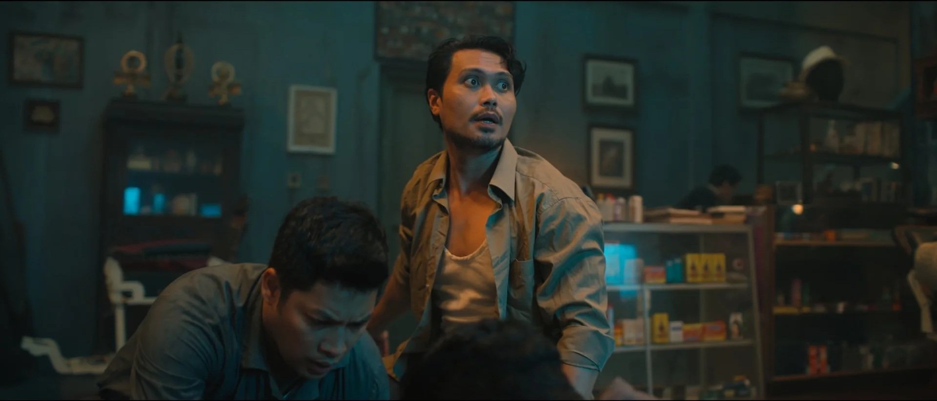 Scene from a movie or show with three men in a dimly lit room, one standing with a surprised expression, two seated looking concerned, shelves with products and pictures on the walls in the background.