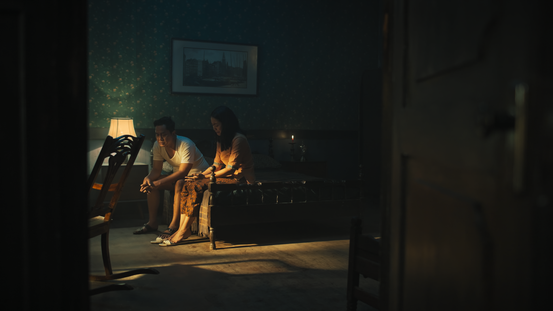 A man and woman sitting on a bed in a dimly lit room, looking down, with a lamp and candles providing soft lighting, and a painting on the wall.