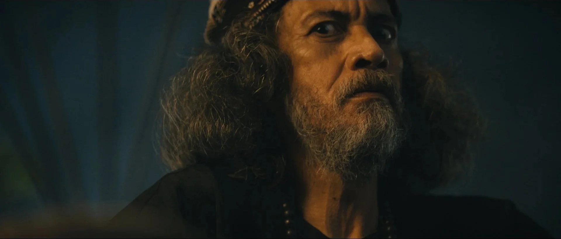 Close-up of an older man with a beard, long curly hair, and a serious expression, wearing a dark outfit, in a dark setting.