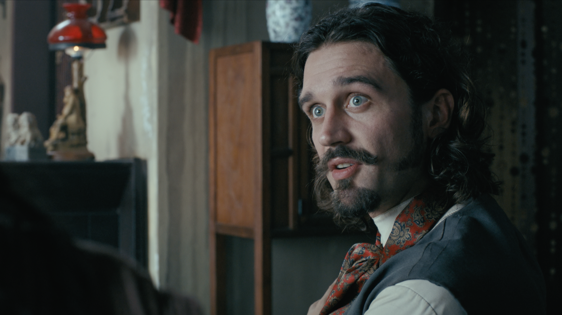 A man with long dark hair, blue eyes, a mustache, and a beard, wearing period clothing with a patterned necktie, gazes to the side in a room with wooden furniture and decor.