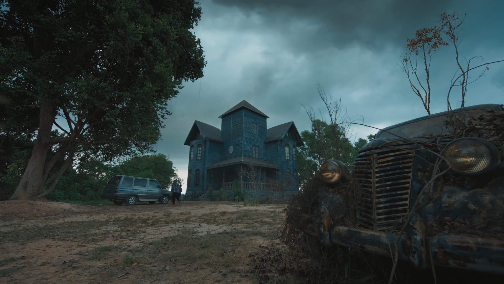 A spooky, old wooden house painted blue in a wooded area, with dark, cloudy skies overhead. An abandoned, rusted car covered with vines and leaves is in the foreground, while a car and a person are seen near the house in the background.
