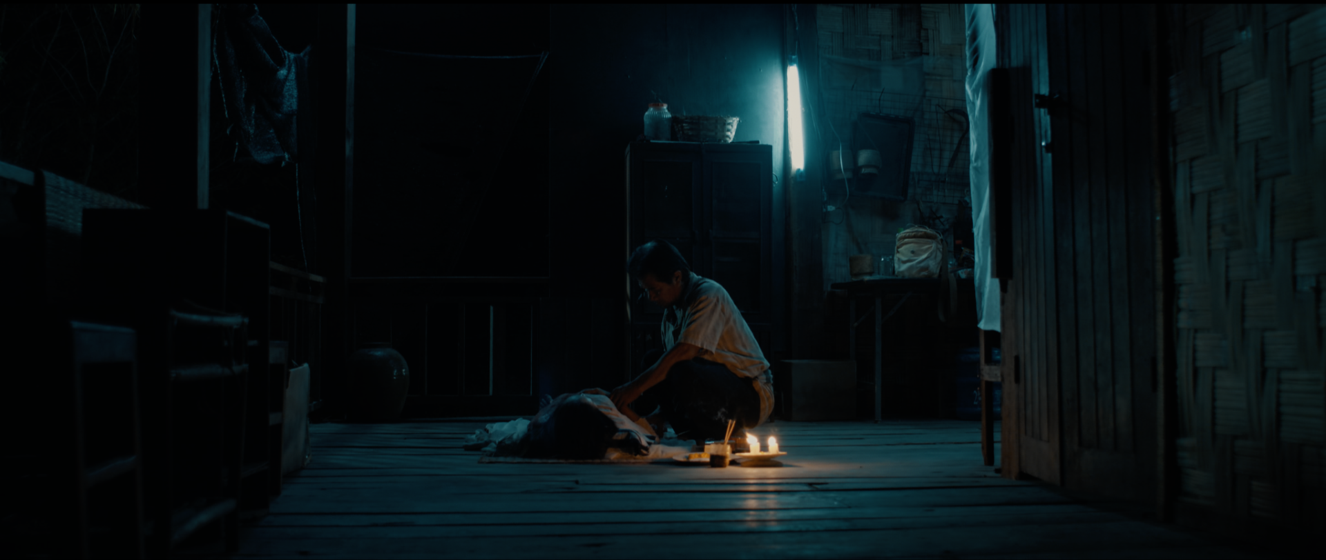A person sitting cross-legged on the floor of a rustic wooden room at night, performing a ritual with candles and incense in front of them. The room is dimly lit and has a blue glow, with furniture and objects scattered around.