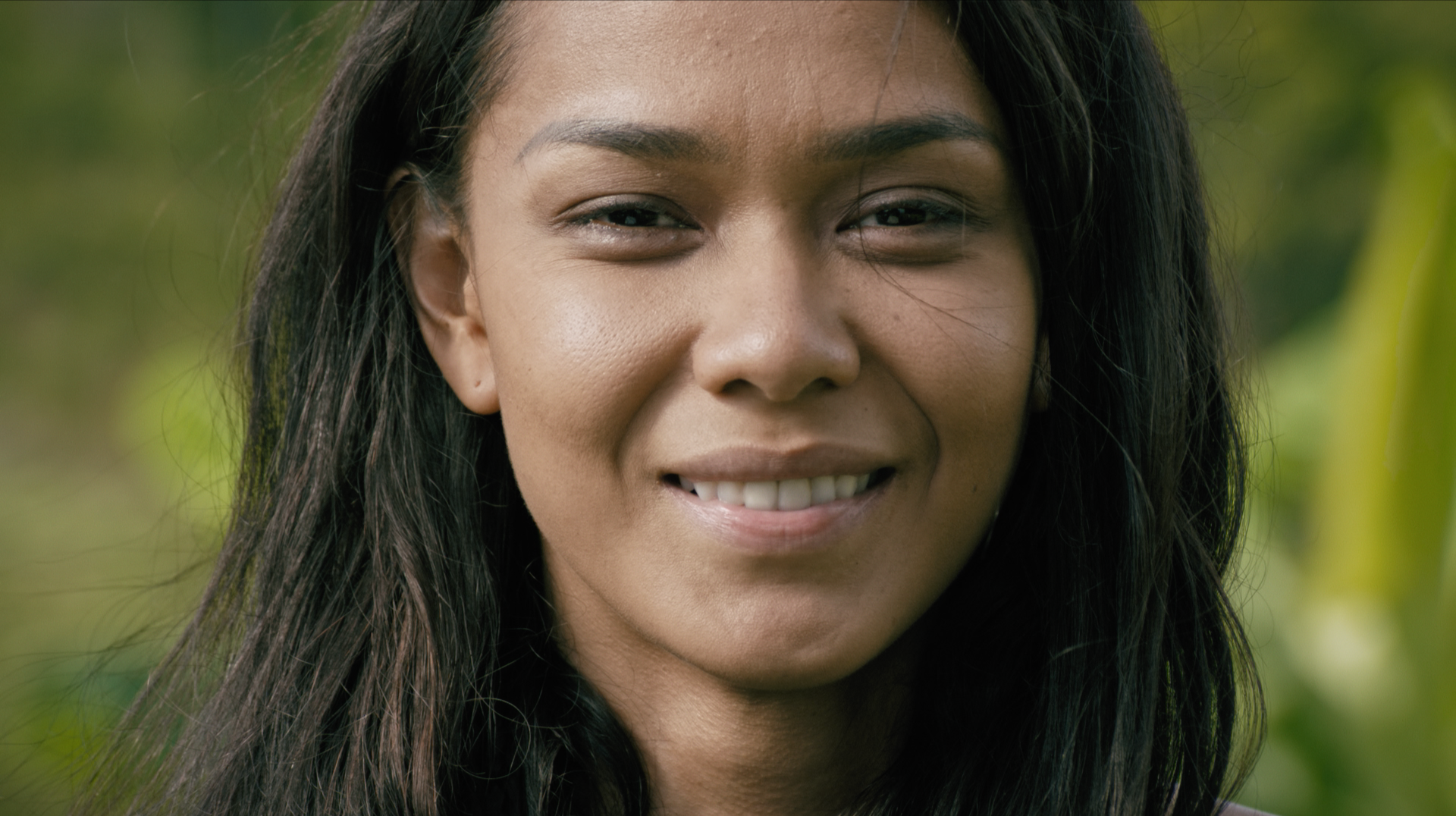 Close-up of a woman with dark hair smiling outdoors with a blurred green background.