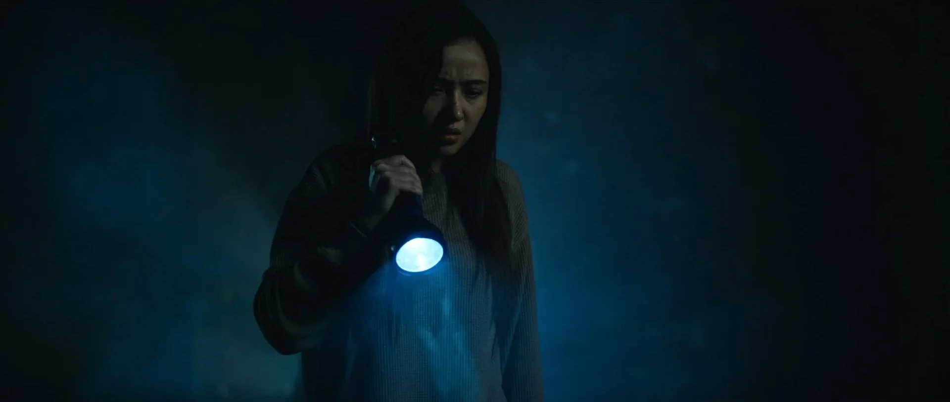 A woman holding a flashlight in a dark, misty environment, looking concerned.