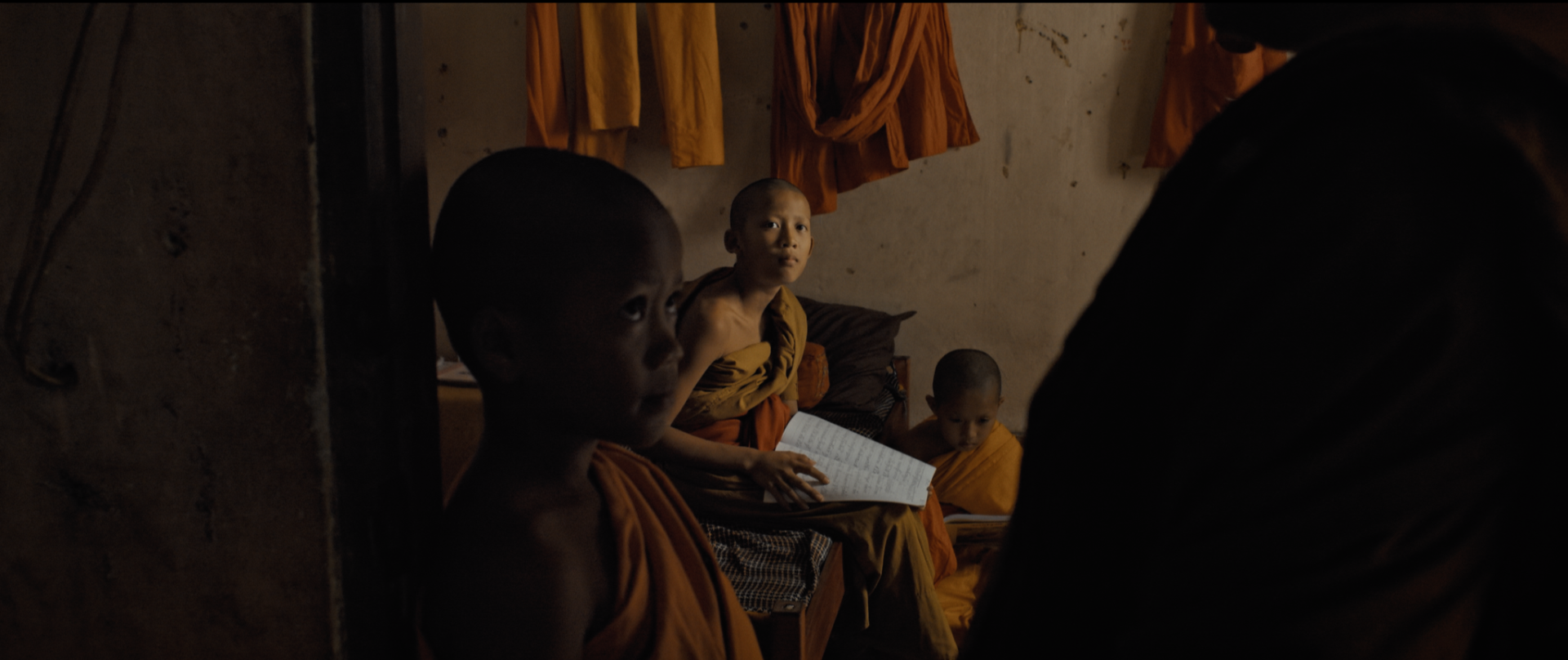 Three young children dressed in orange robes sit inside a dimly lit room, with one of them holding an open book, and a woman sitting across from them. The background shows hanging orange cloths and a concrete wall.