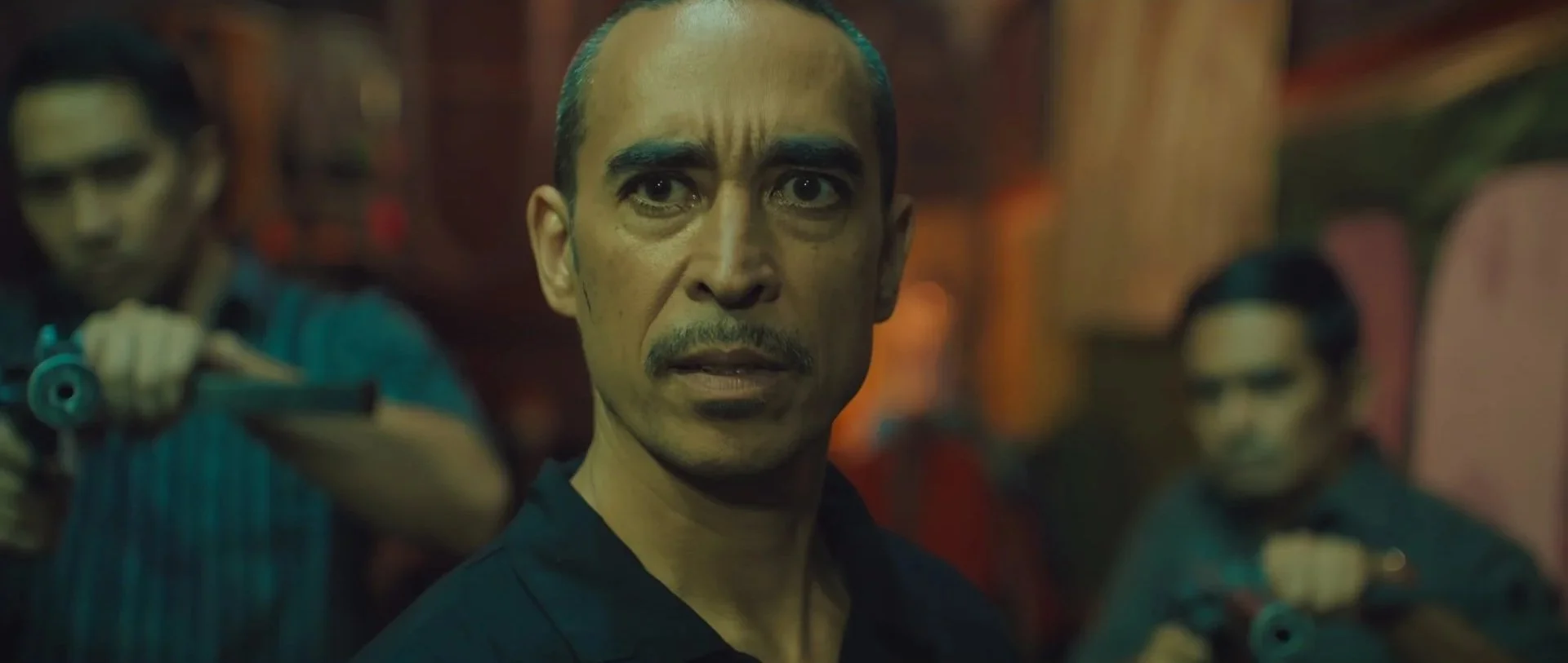 A man with a serious expression faces the camera, surrounded by other people, in a dimly-lit environment with warm lighting.