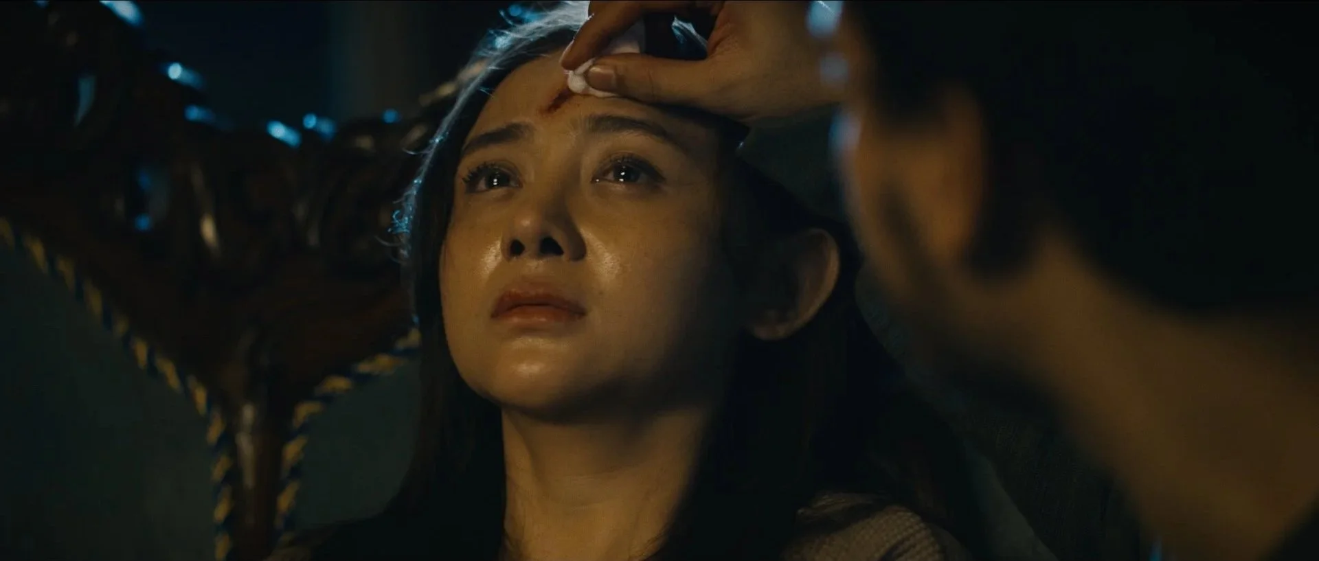 A woman lying down with a worried expression, someone is wiping a bloody wound on her forehead.