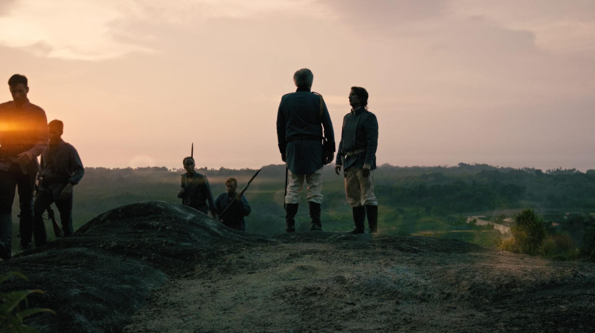 Silhouettes of people standing on a rocky hill at sunset, some holding rifles, with a distant landscape in the background.