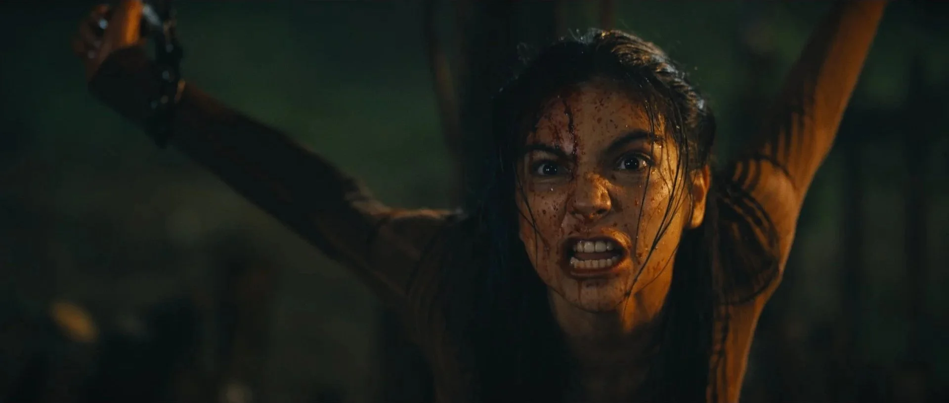 A woman with blood on her face and torn clothing looks intensely forward with a fierce expression.