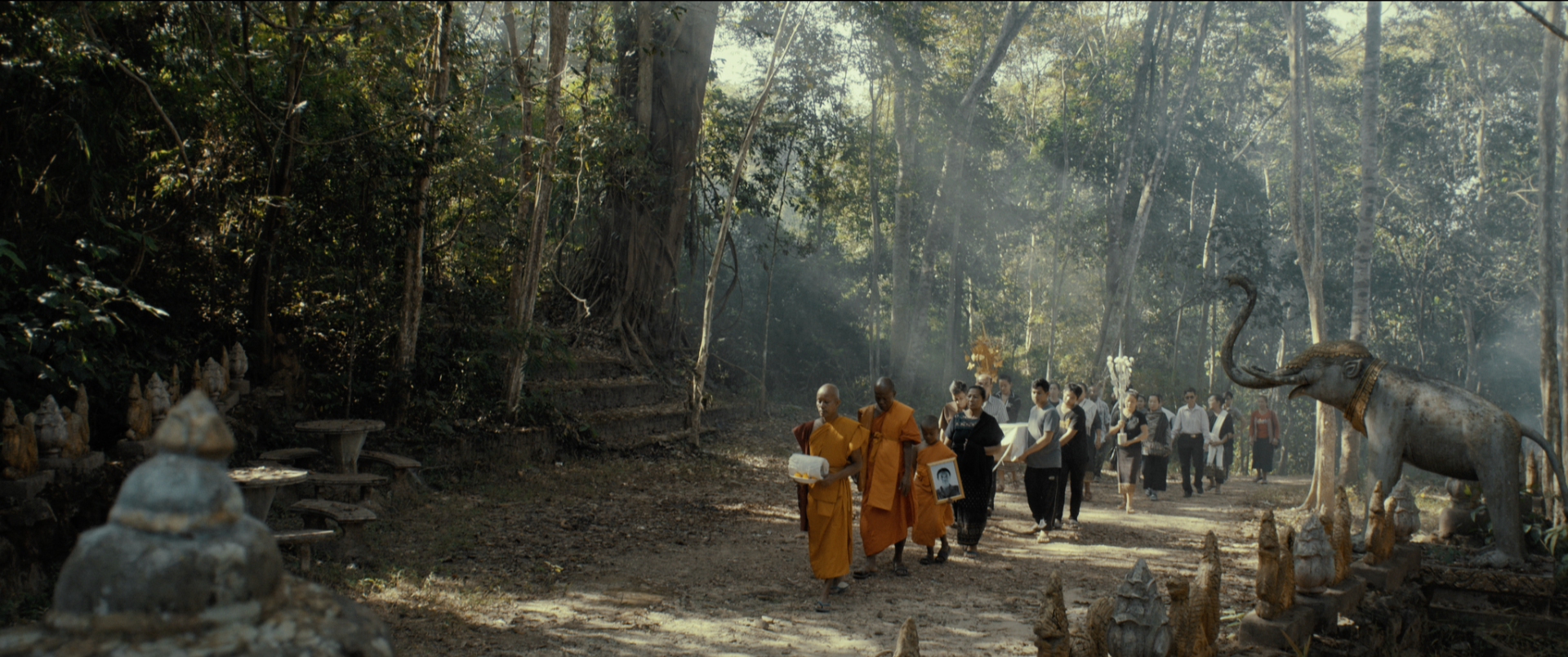 A group of monks and laypeople walking through a forested area during a funeral procession, carrying a framed photograph and floral arrangements, with statues and a large elephant statue nearby.
