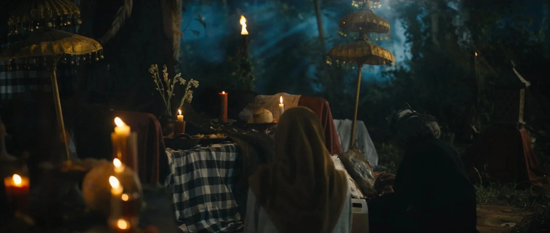 Slightly illuminated outdoor nighttime scene with tables, candles, and umbrellas, people sitting and standing, surrounded by trees and foliage.