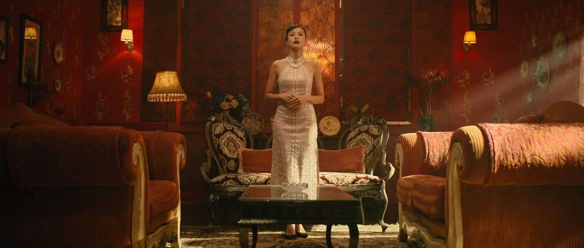A woman in a long, elegant gown standing in a vintage-styled living room with ornate furniture, warm lighting, and decorative wall art.