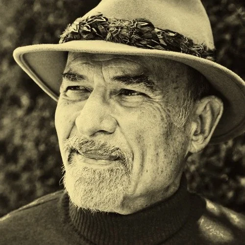 A close-up of an elderly man wearing a wide-brimmed hat with a decorative band, outdoors in natural light.