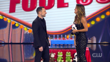 A man and woman stand on stage, smiling and talking. The woman holds a soccer ball. Behind them is a large birthday cake with candles and colorful decorations, including stuffed animals and a 'FOLIE S' sign.