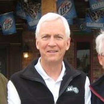 Older man with white hair wearing a black North Face vest and white shirt, standing outdoors near a Miller Lite sign.