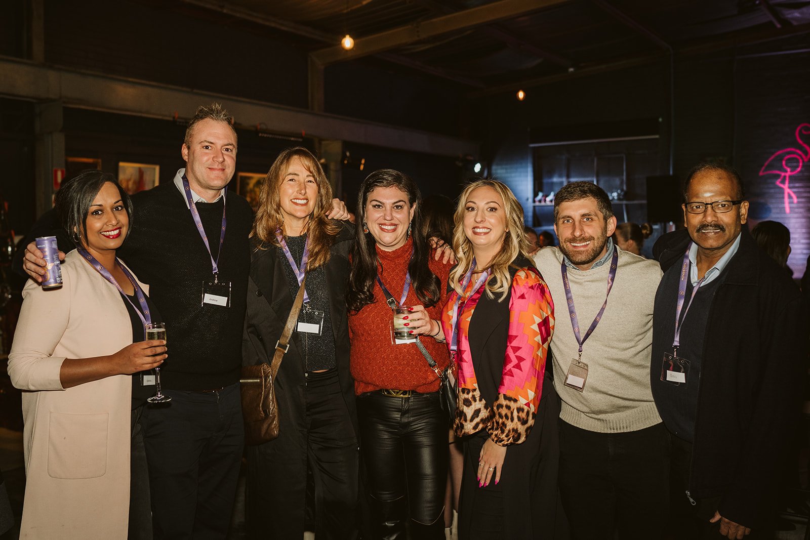 A diverse group of seven people enjoying an indoor gathering, wearing lanyards, and smiling at the camera. Some are holding drinks, and the setting has a casual, friendly atmosphere.