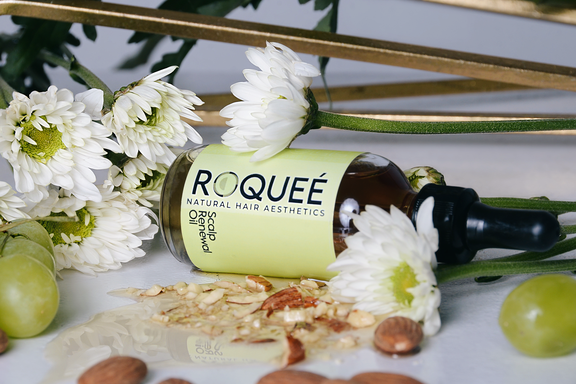 A bottle of Roqué natural hair aesthetics scalp renewal oil laying on its side with white daisies and scattered nuts and seeds around it.