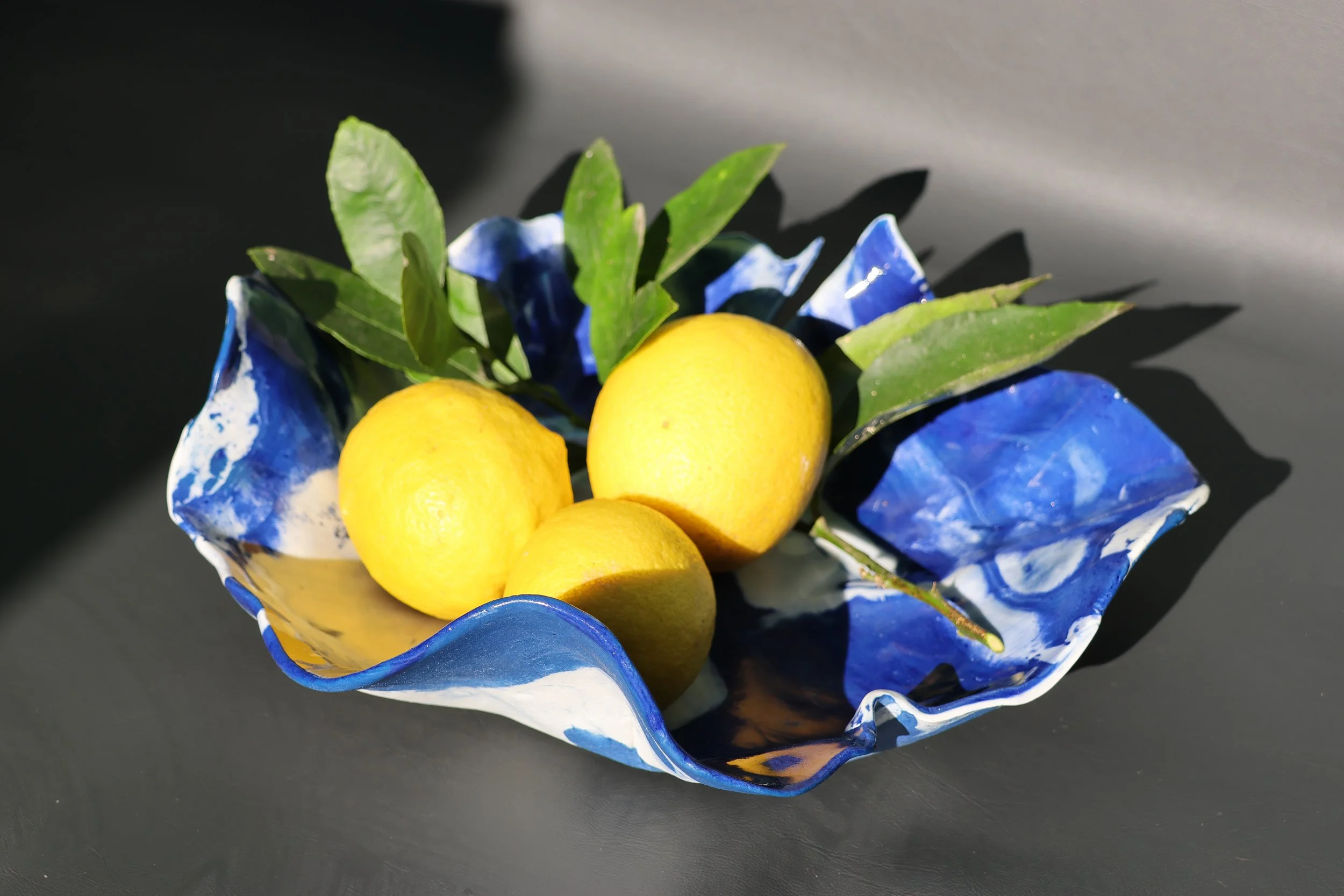 A decorative bowl with a blue and white marbled pattern, holding three yellow lemons and some green leaves, set on a dark surface.