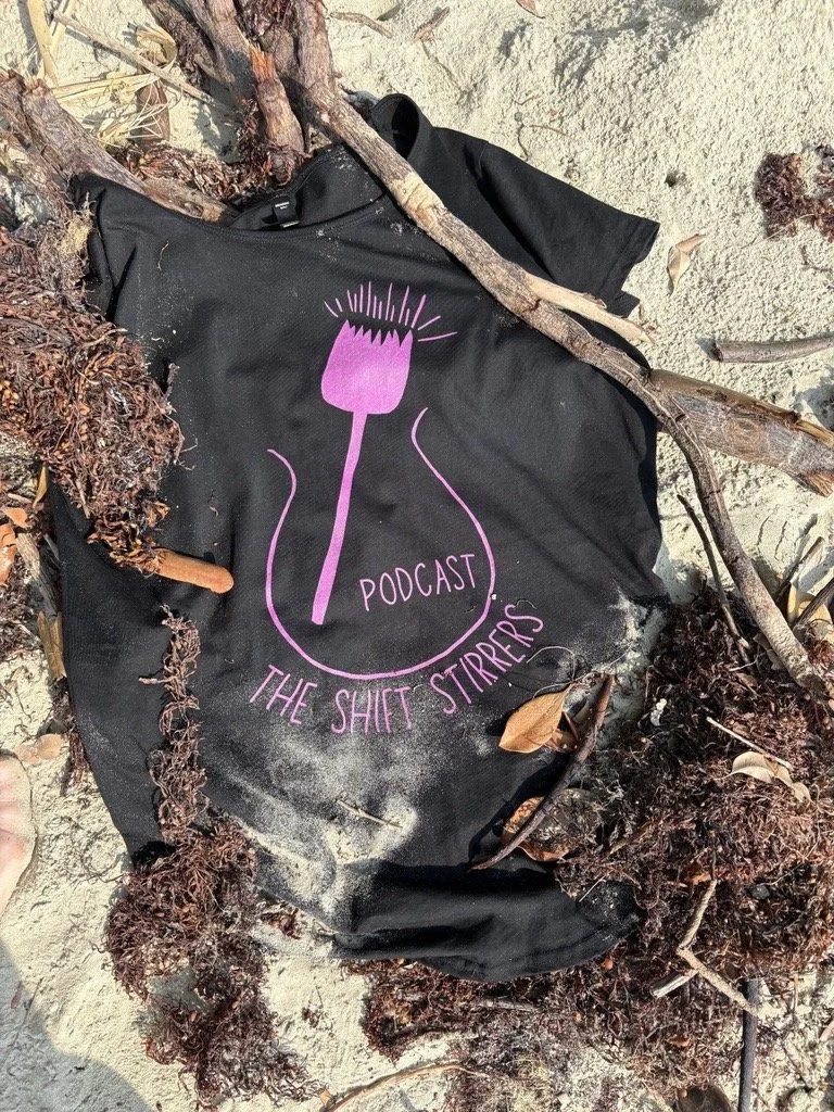 Black tote bag with pink logo and text, lying on sandy ground surrounded by twigs, leaves, and seaweed.