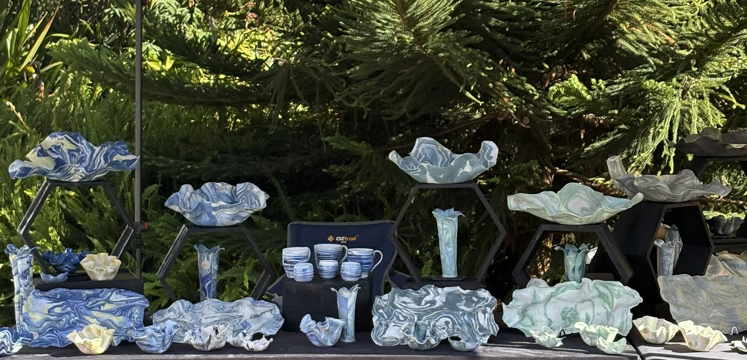 Michellè Benson Ceramics | Display of handmade marbled pottery bowls, vases, and cups on black shelves outdoors, with green foliage in the background.