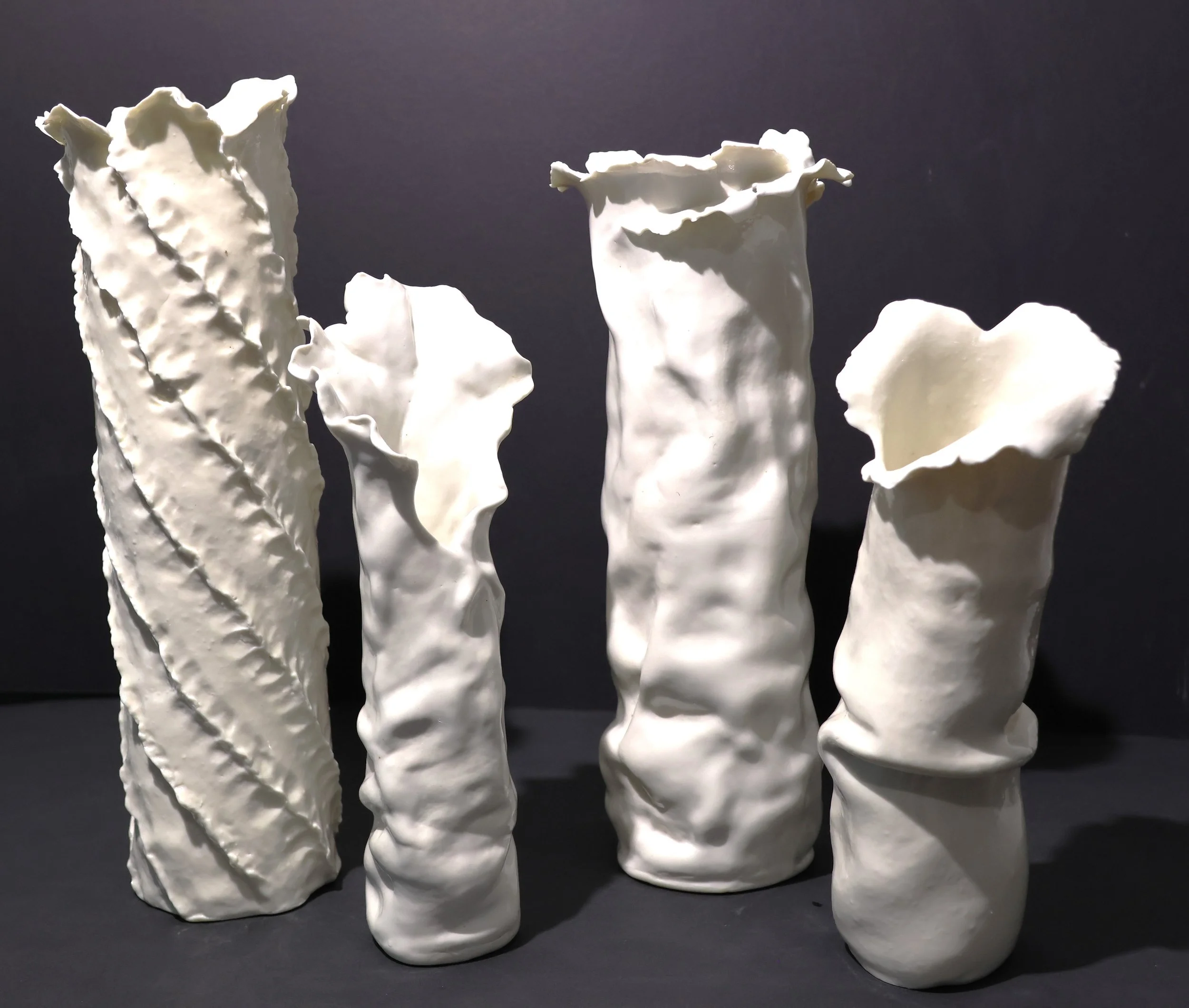 Four white ceramic vases with uneven, textured surfaces and irregular, ruffled openings, arranged on a black surface against a dark background.