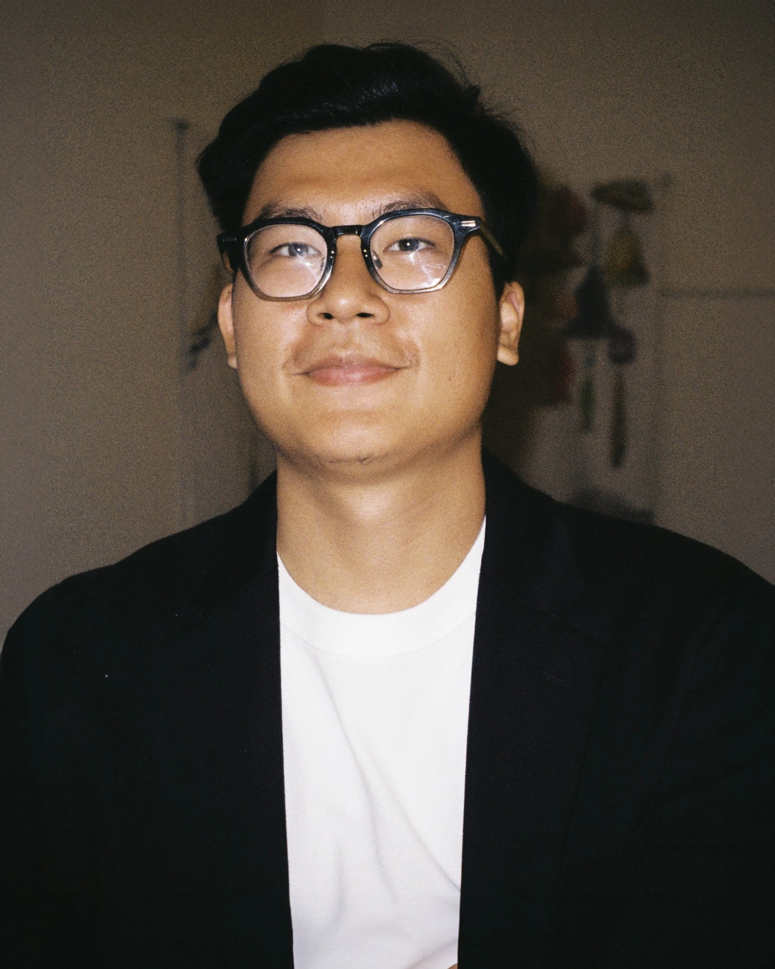 Young man with black hair and glasses smiling in a black jacket and white shirt.