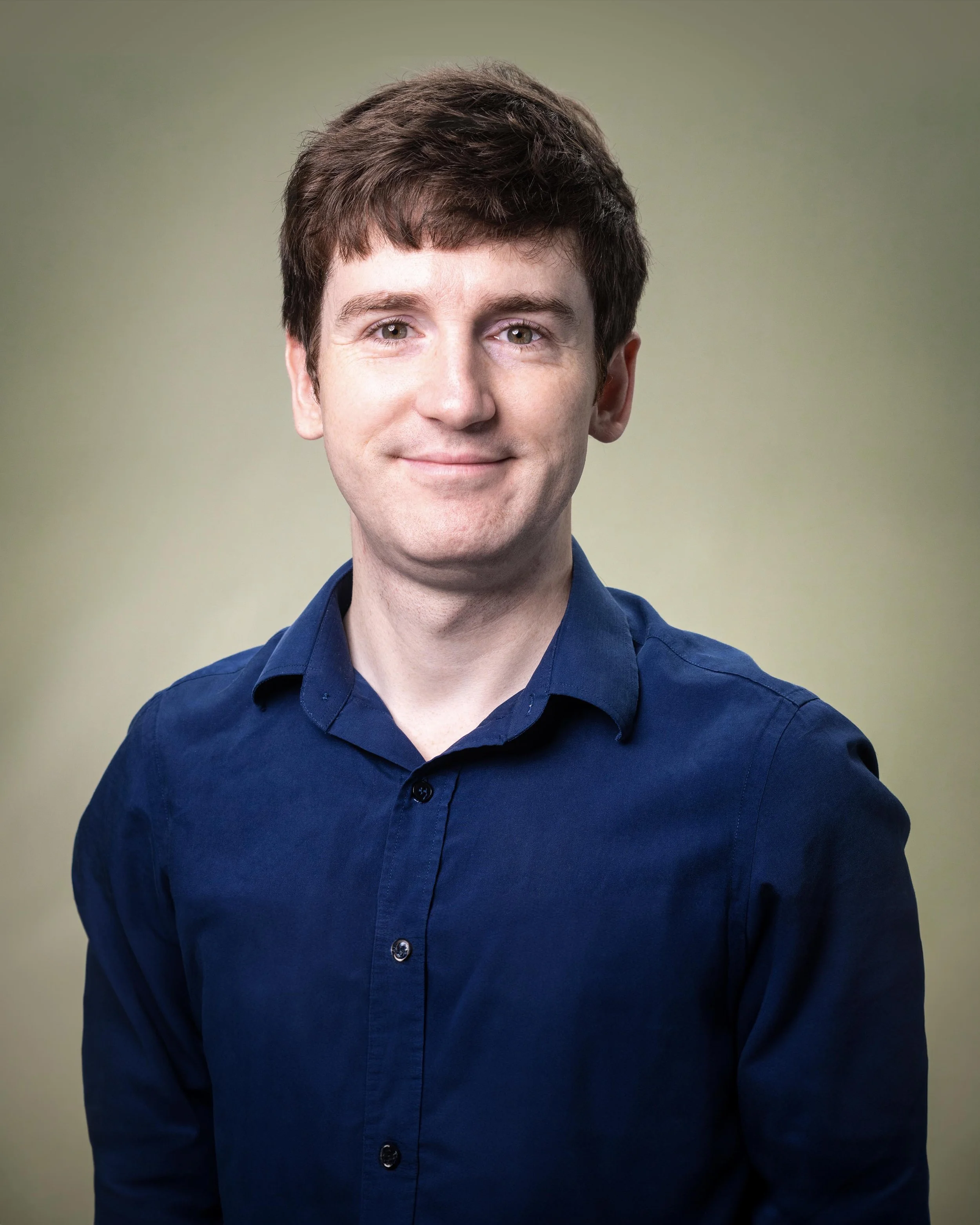 A man with brown hair, wearing a navy blue button-up shirt, smiling at the camera against a light green background.