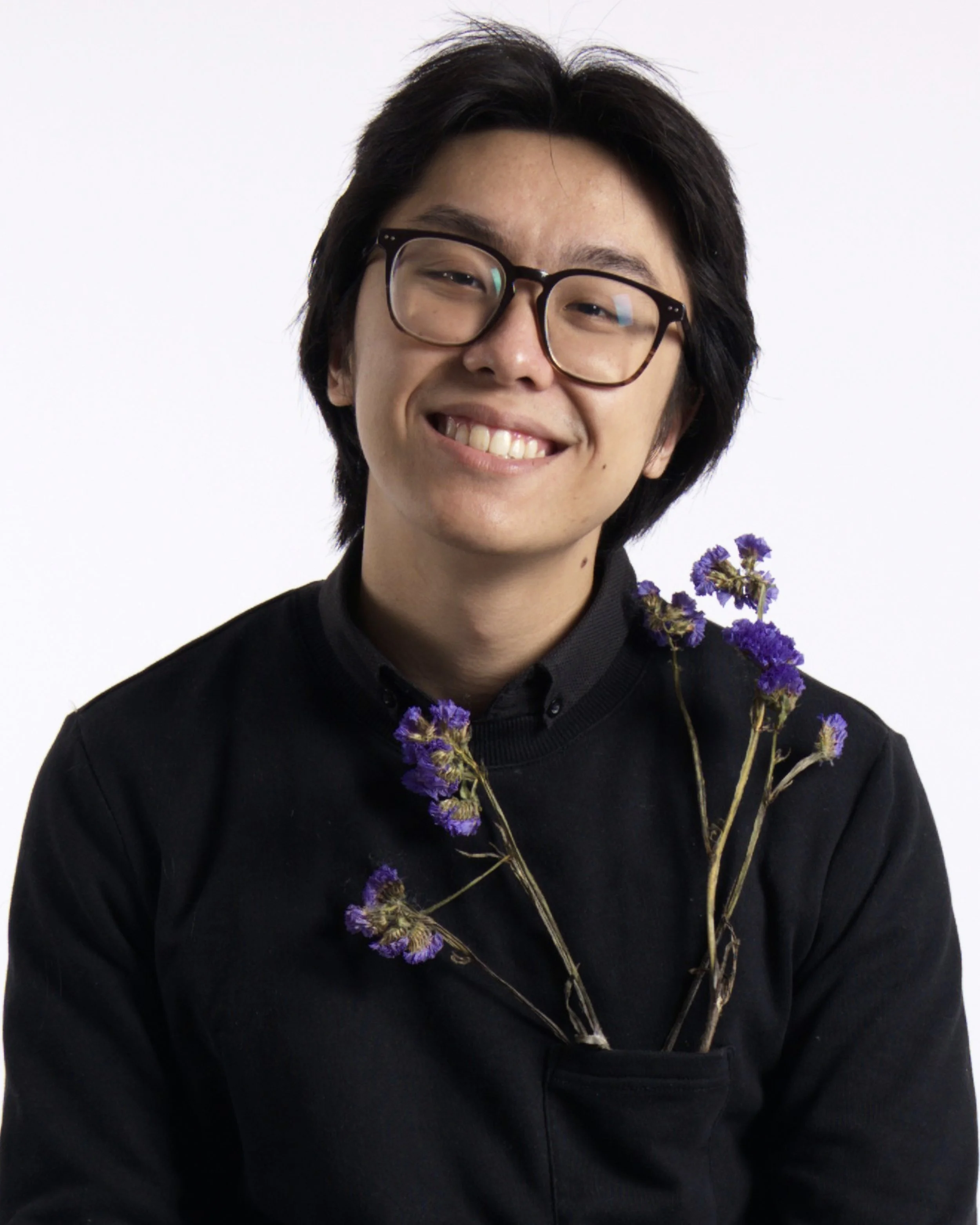 Young man with glasses smiling, wearing a black shirt and holding purple flowers in front of a white background.