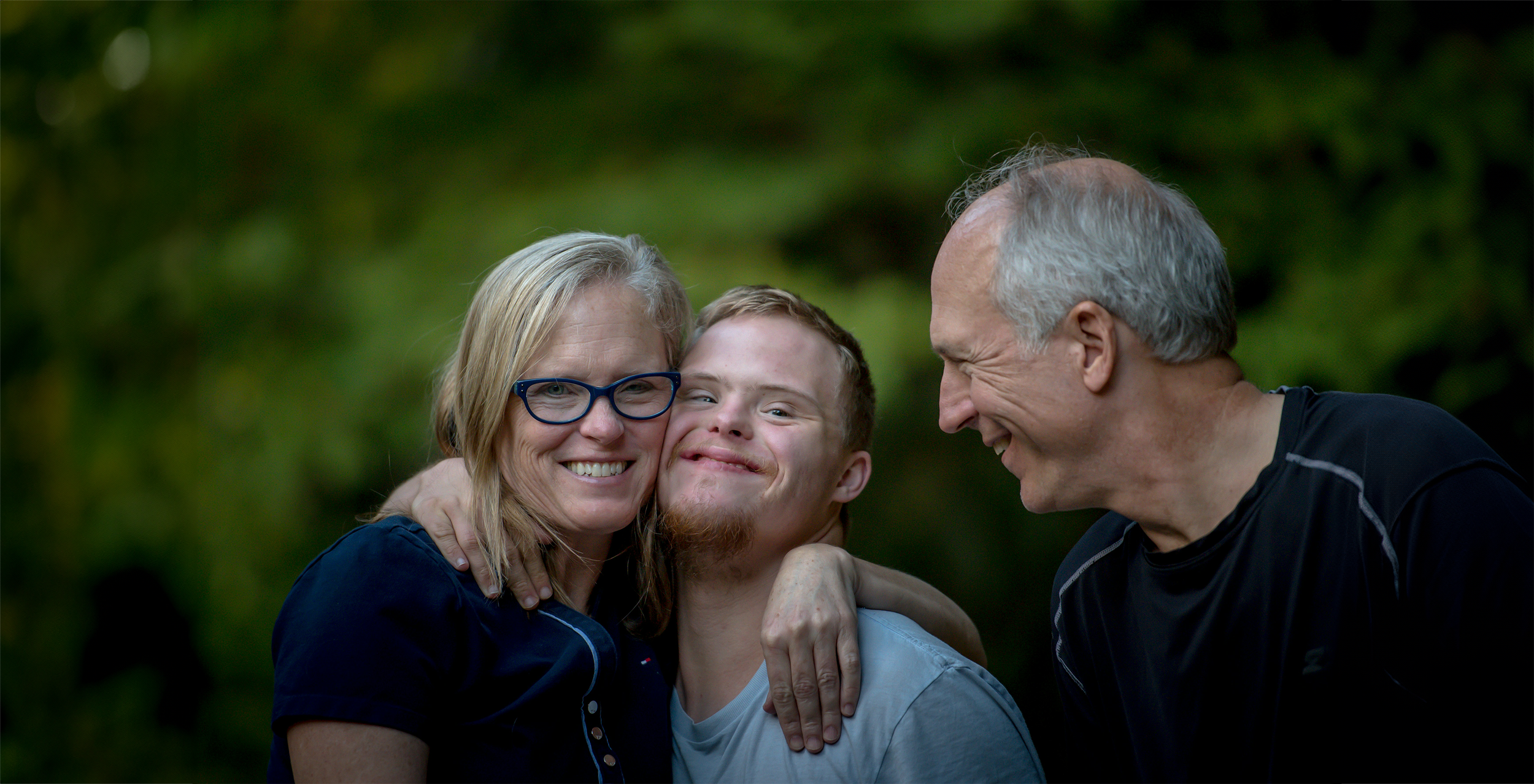 A happy family of three hugging outdoors with a green, leafy background.